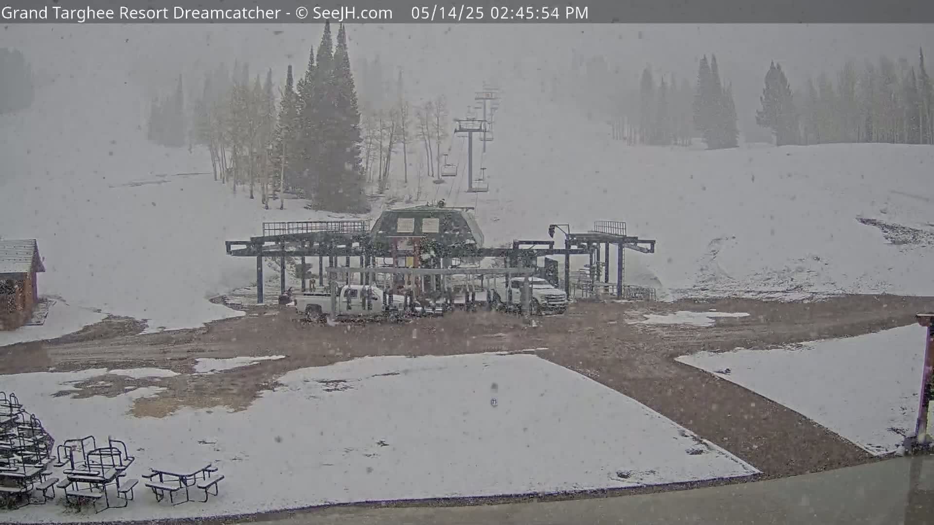 A snow-covered ski lift base area with several vehicles and picnic tables is seen during a light snowfall.