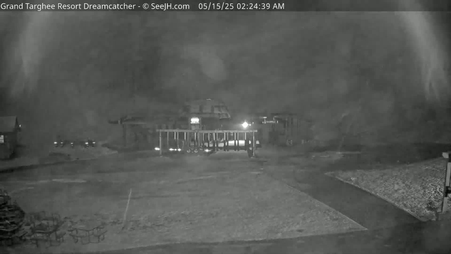A nighttime, foggy view shows a gate and several vehicles parked near a building.