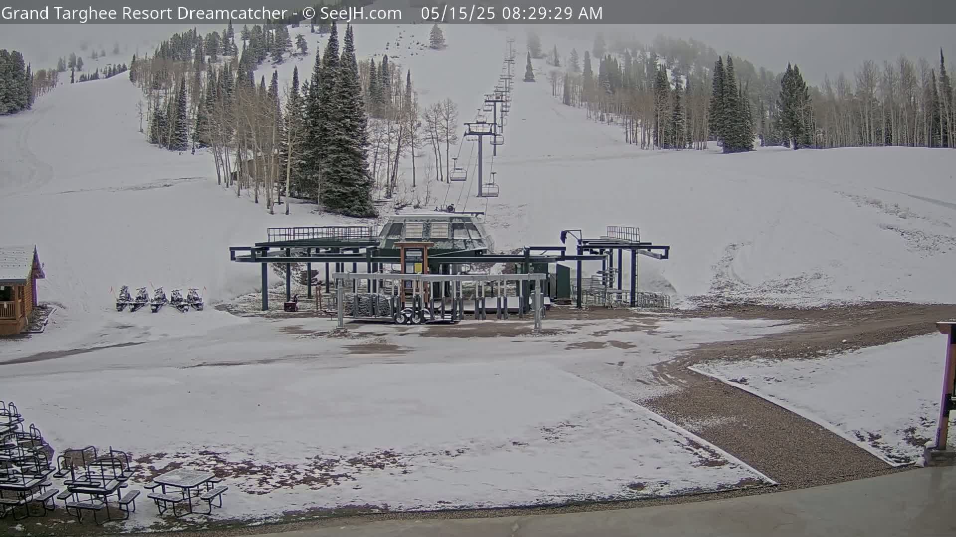 A snow-covered ski resort base area with a chairlift, several snowmobiles, and picnic tables is visible under an overcast sky.