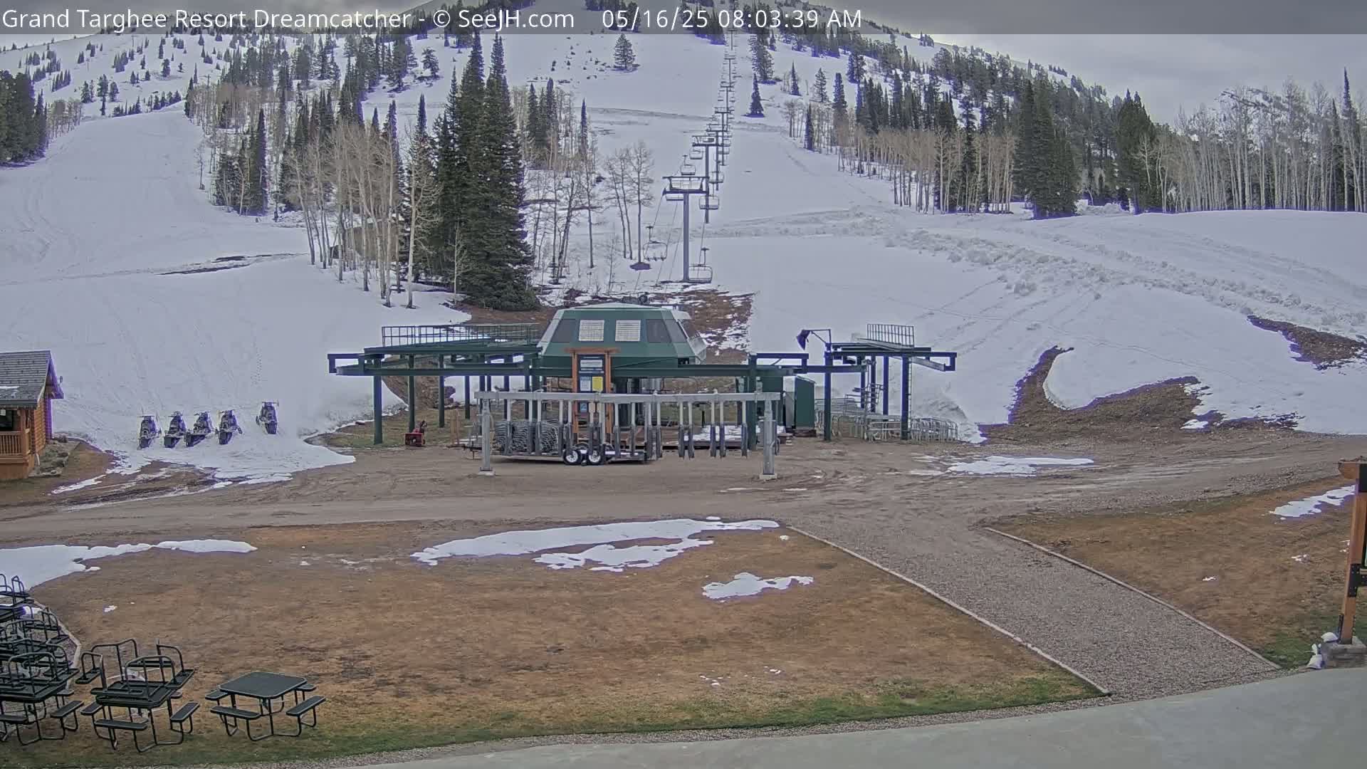 A snow-covered ski slope with a chairlift and several snowmobiles parked near a base lodge, under overcast skies.