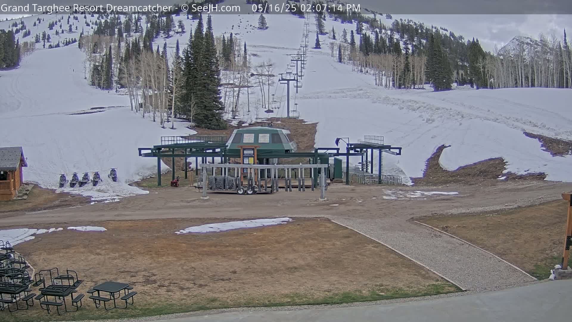 A ski lift base station sits in a mostly snow-free area at the base of a snow-covered mountainside under an overcast sky.