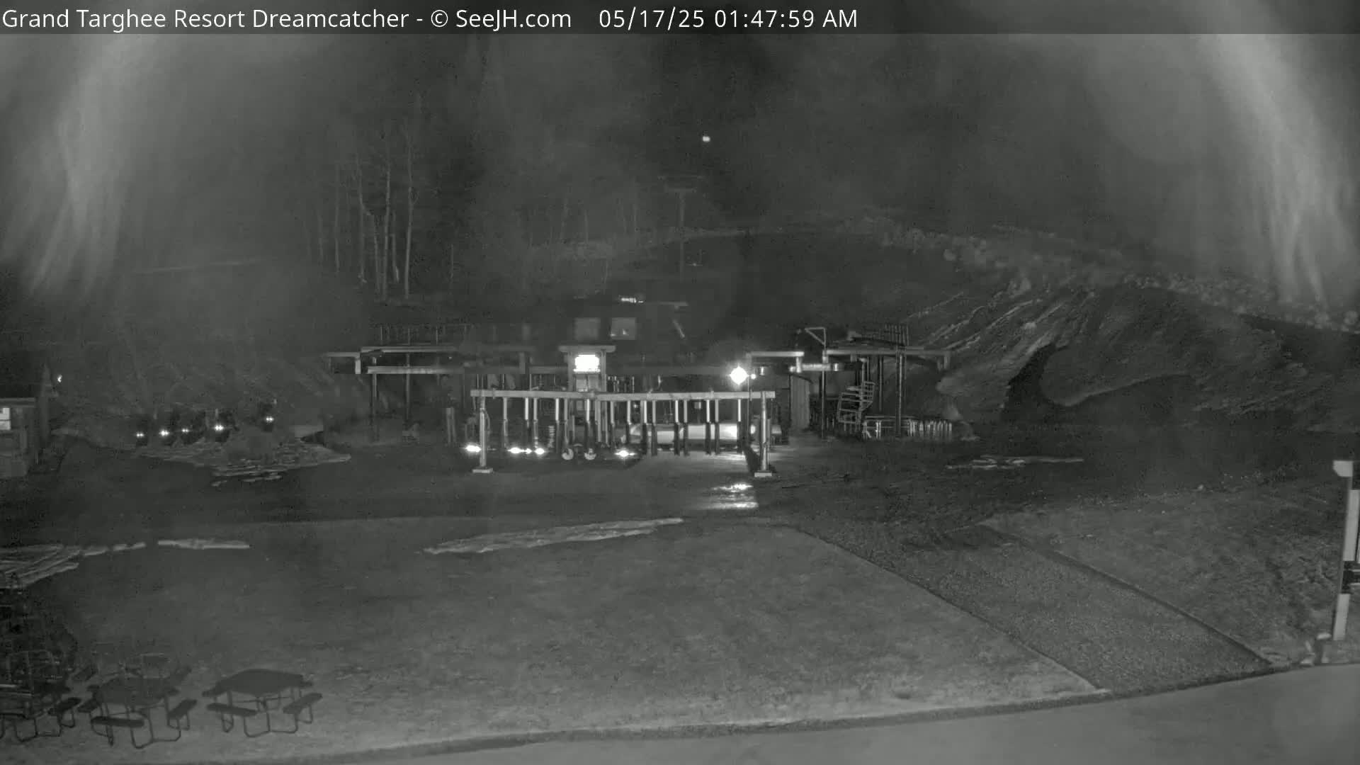 A nighttime, low-light image shows a gated area with several vehicles parked inside, picnic tables in the foreground, and a dark, hilly landscape in the background.