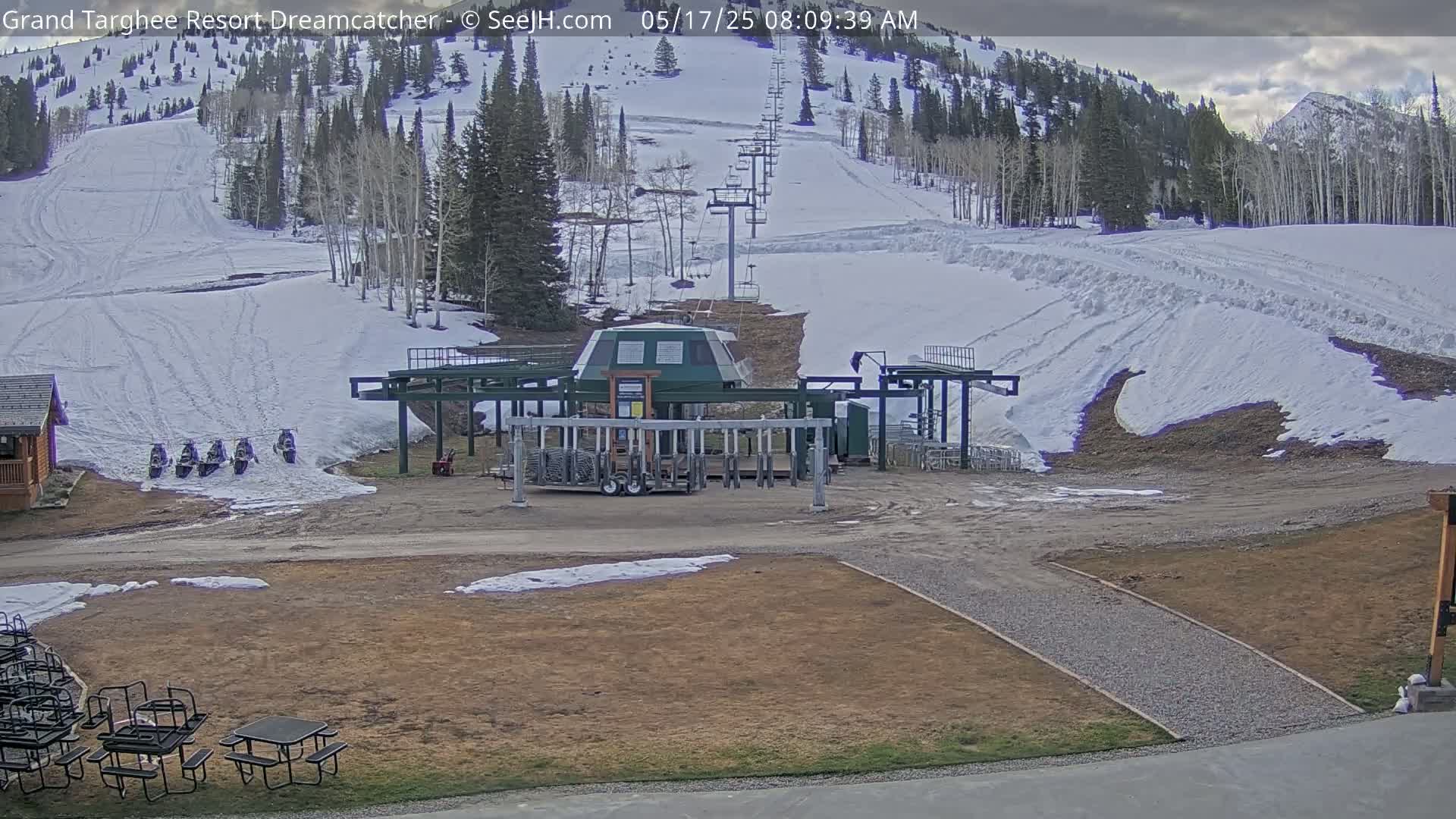 A ski lift base area with snow-covered slopes in the background, under cloudy skies.