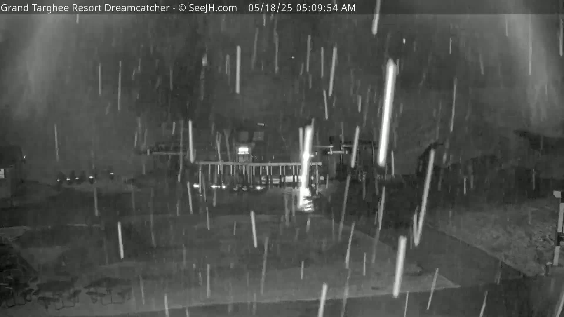 A nighttime, snowy scene shows a building with a fence and patio furniture in the foreground.