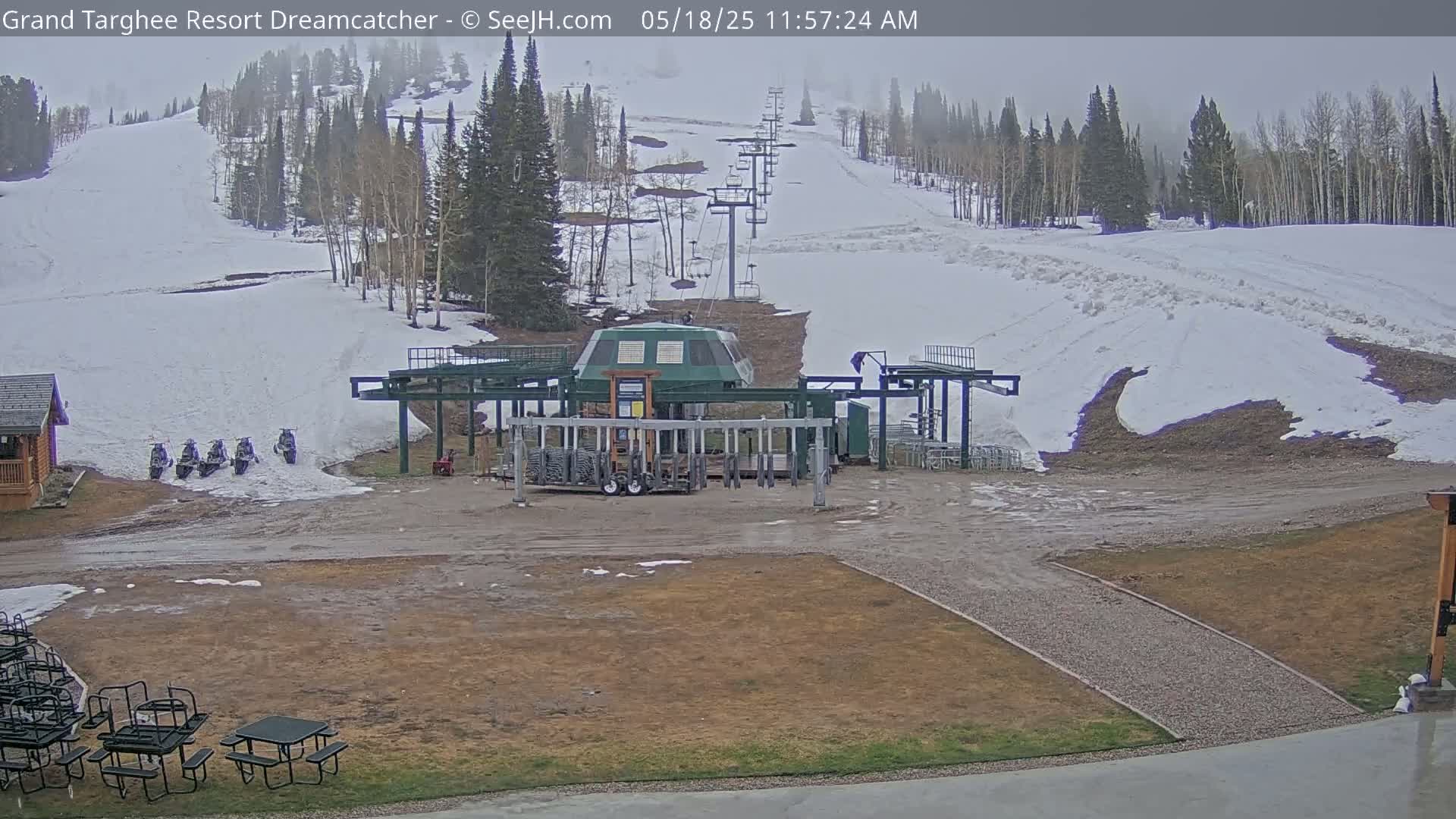 A ski lift base station sits in a muddy area next to a snow-covered ski slope on an overcast day.