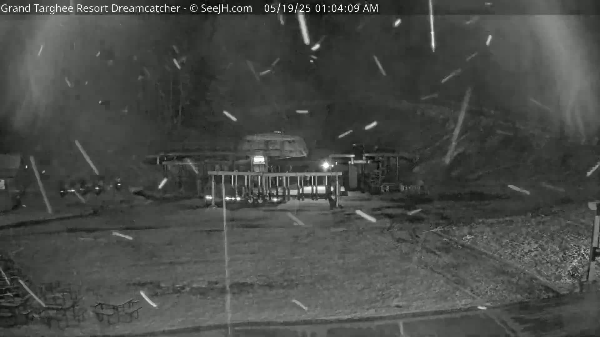 A nighttime, snowy scene shows a large structure with a gate, and several rows of empty chairs in the foreground.