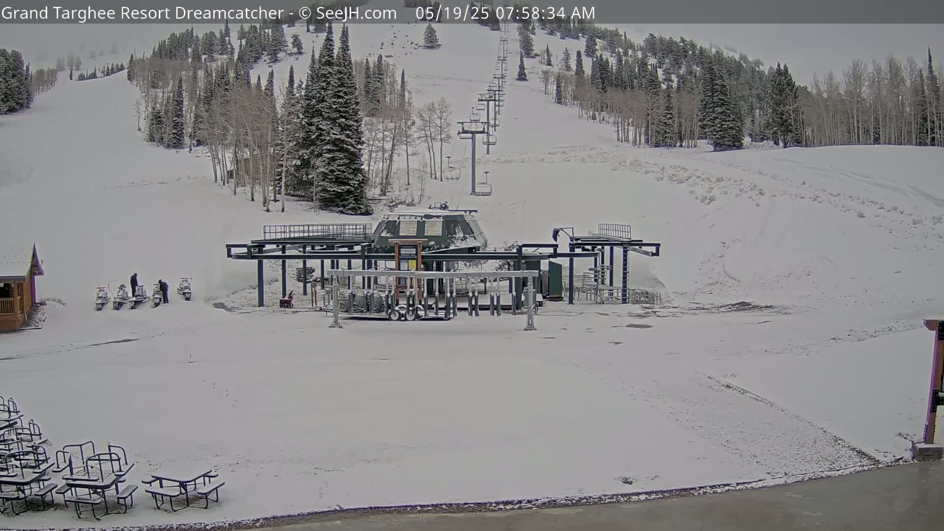 A snow-covered ski lift base area with a chairlift ascending a snow-covered hill, several snowmobiles parked nearby, and picnic tables scattered in the foreground.
