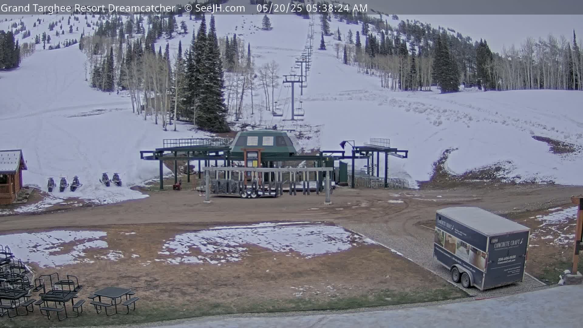 A ski lift base area with snow on the ground and a chairlift ascending a snow-covered mountainside under an overcast sky.