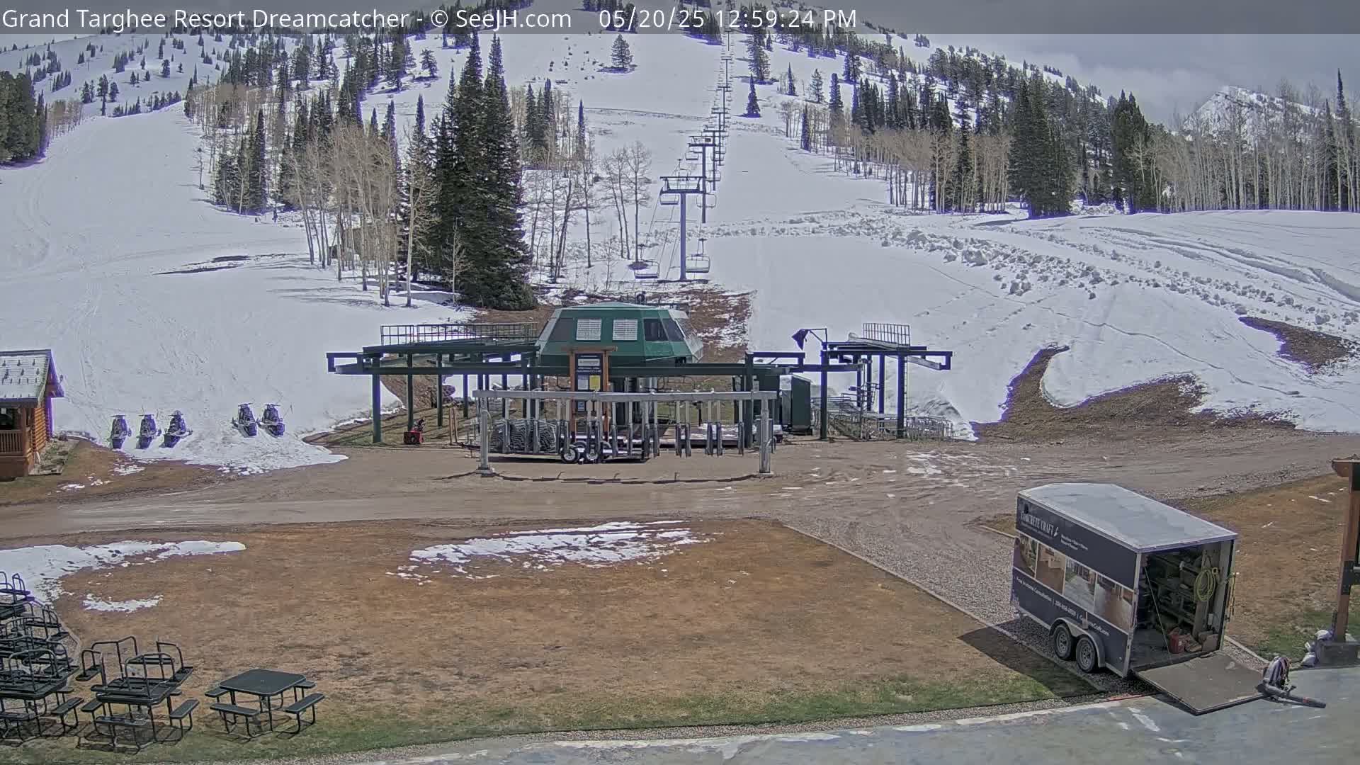A ski lift base station sits in a partially snow-covered area, with a mountainside of trees and snow visible in the background under partly cloudy skies.