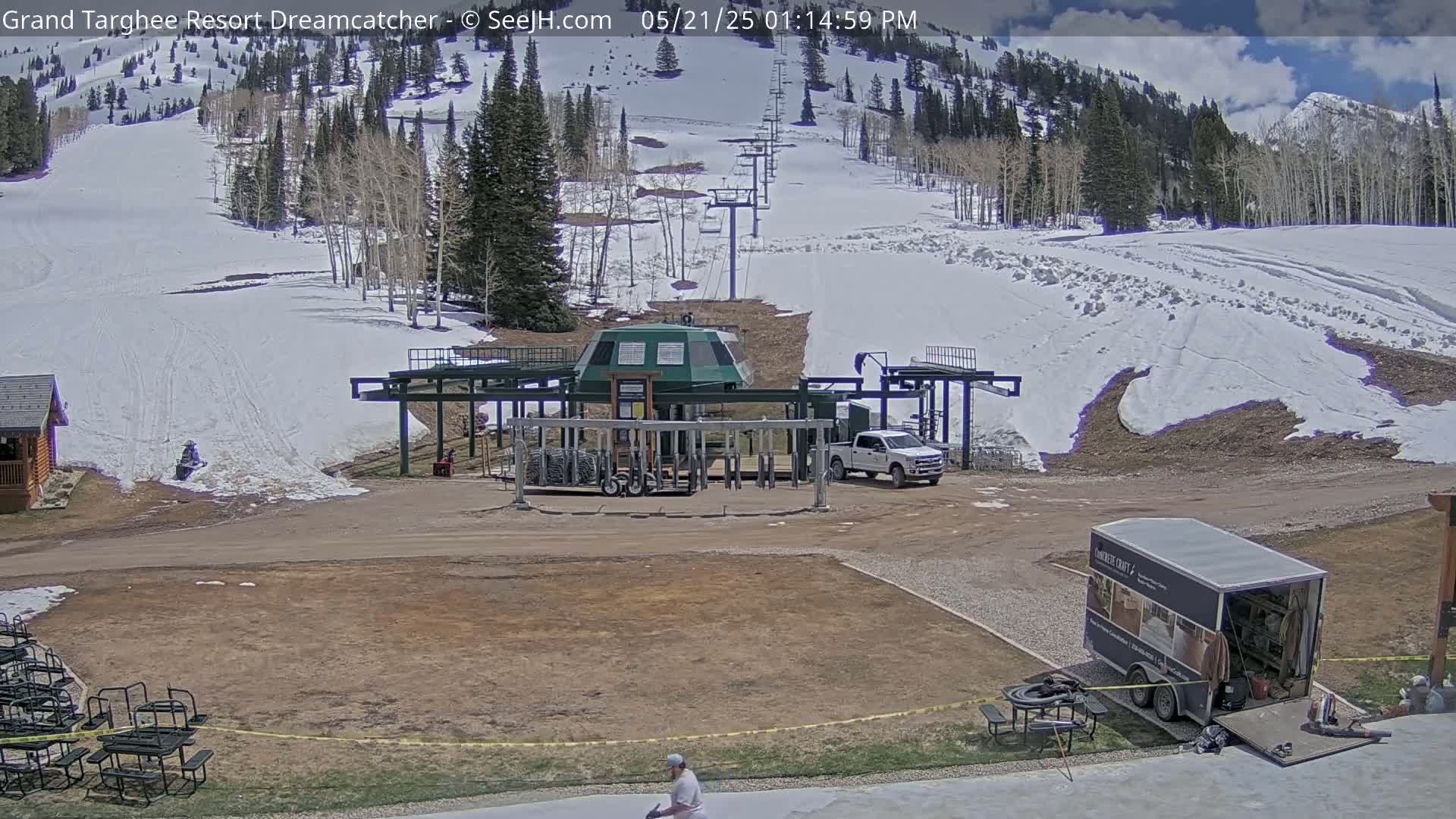 A mostly sunny day shows a ski lift base area with some remaining snow on the slopes, a pickup truck, and a trailer.