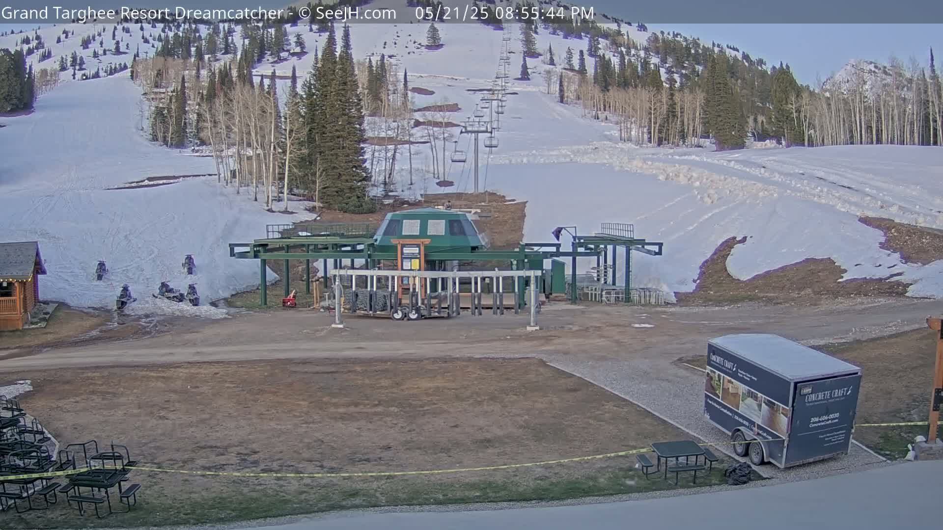 A ski lift base station sits in a snowy mountain landscape with several snowmobiles parked nearby under a clear sky.