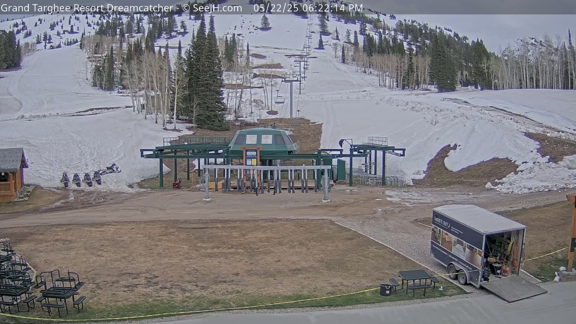 A ski lift base station sits in a mostly bare, snow-dusted landscape under an overcast sky, with several snowmobiles parked nearby and a truck parked further away.