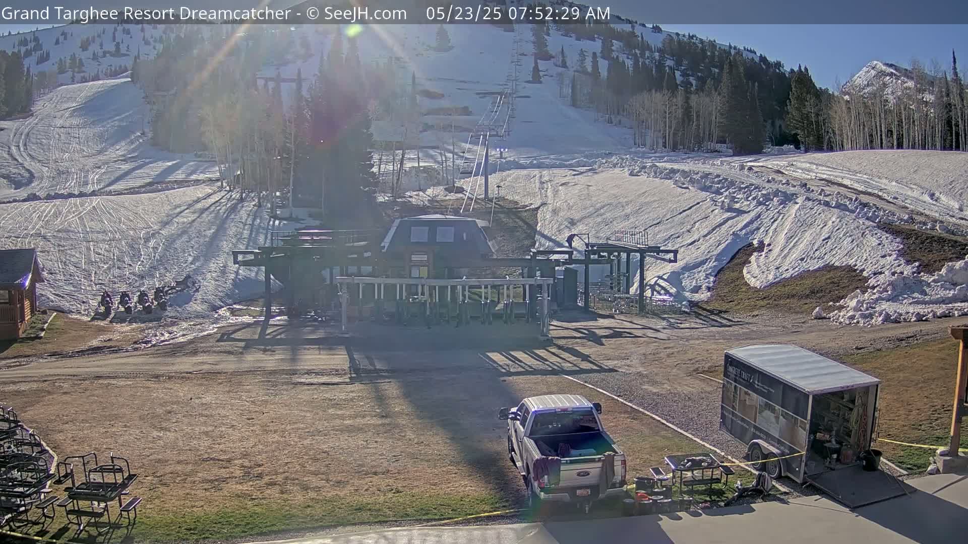 A ski lift base area with a pickup truck and trailer is visible under sunny skies, with snow still present on the surrounding slopes and hills.