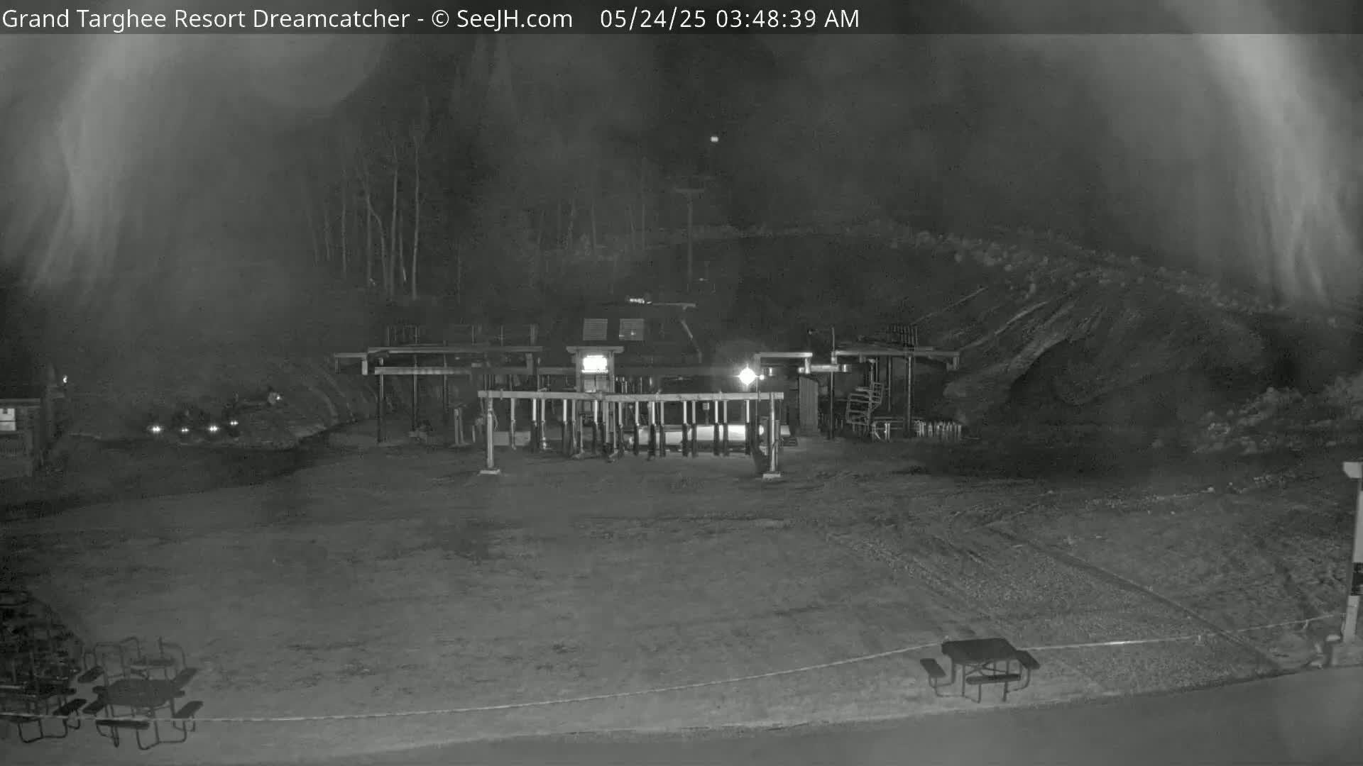 A nighttime, low-light view shows a mostly empty outdoor area with picnic tables, a vehicle at a gated entrance, and a hazy atmosphere.