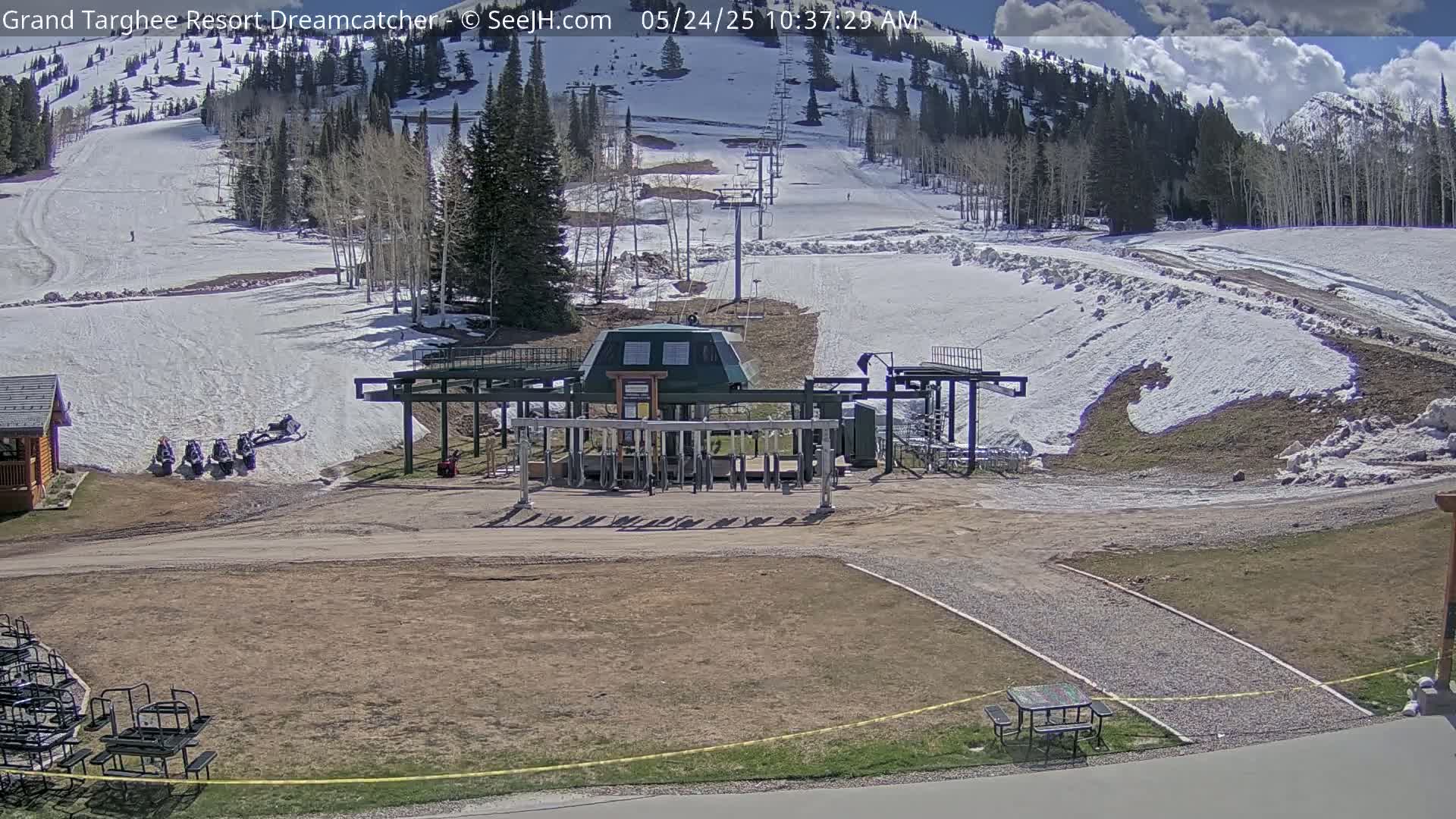 A ski lift base station sits on a mostly bare, dirt ground with patches of melting snow in the foreground and a snow-covered mountainside in the background under a partly sunny sky.