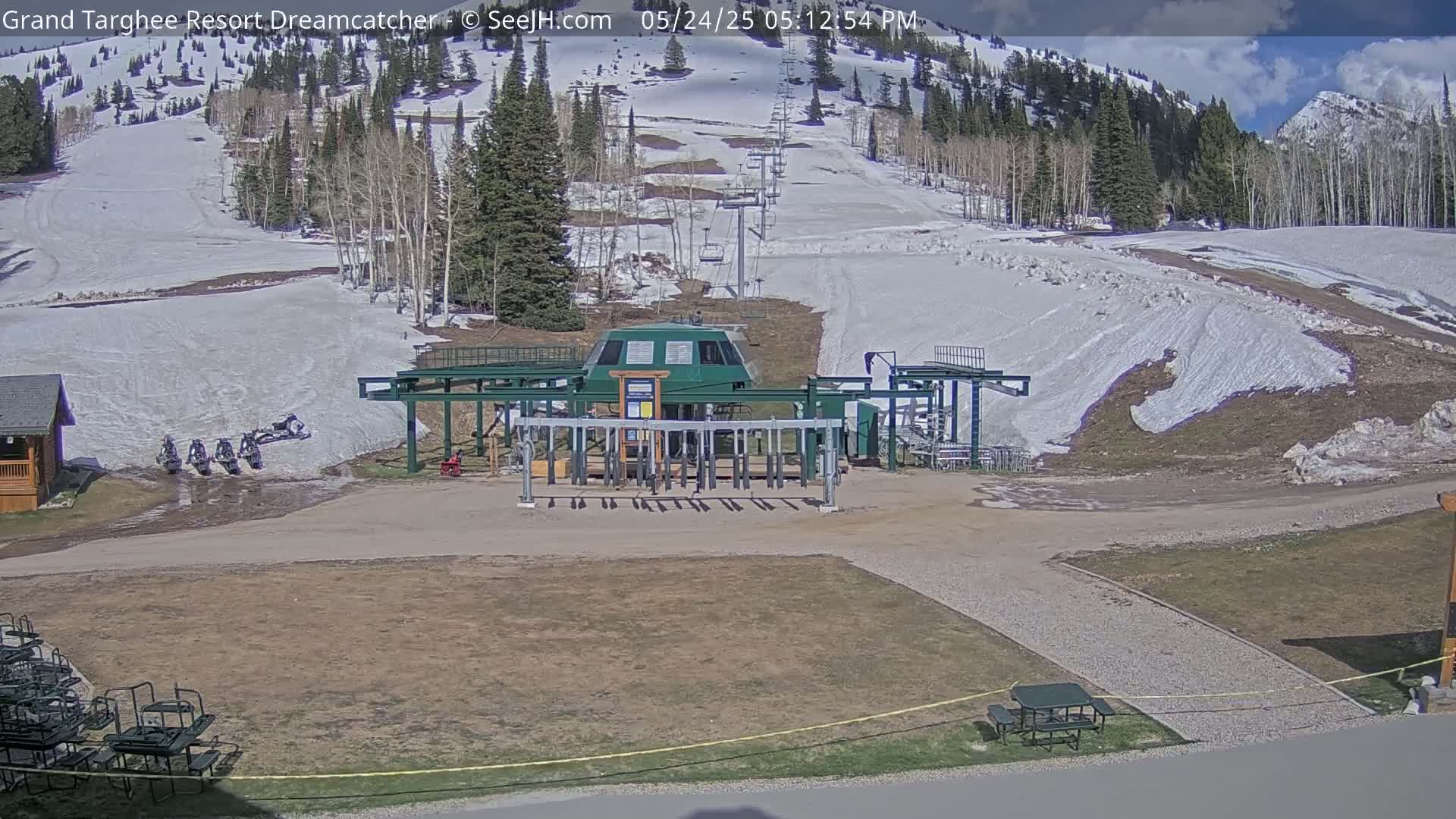 A ski lift base station sits in a sunny, partially snow-covered mountain landscape.