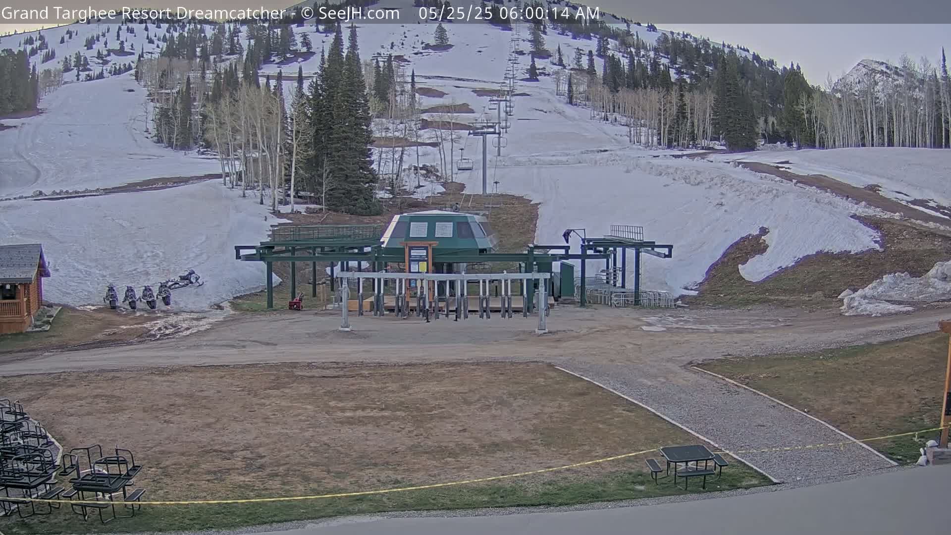 A ski lift base station sits at the bottom of a snow-covered ski slope, with several snowmobiles parked nearby under a partly cloudy sky.