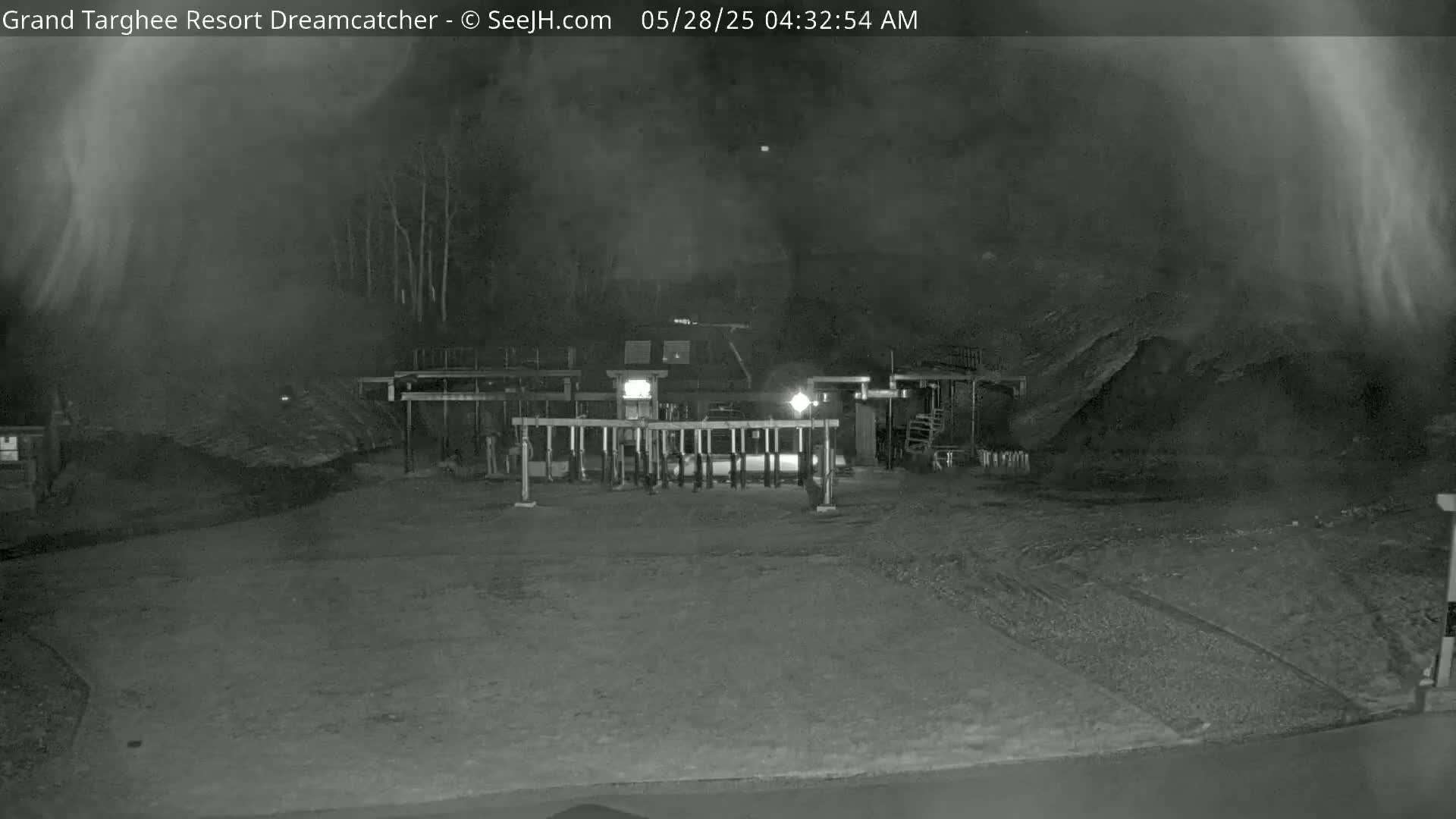 A nighttime, low-light view of a mostly empty parking lot with a gate and a building in the background, under hazy or foggy conditions.