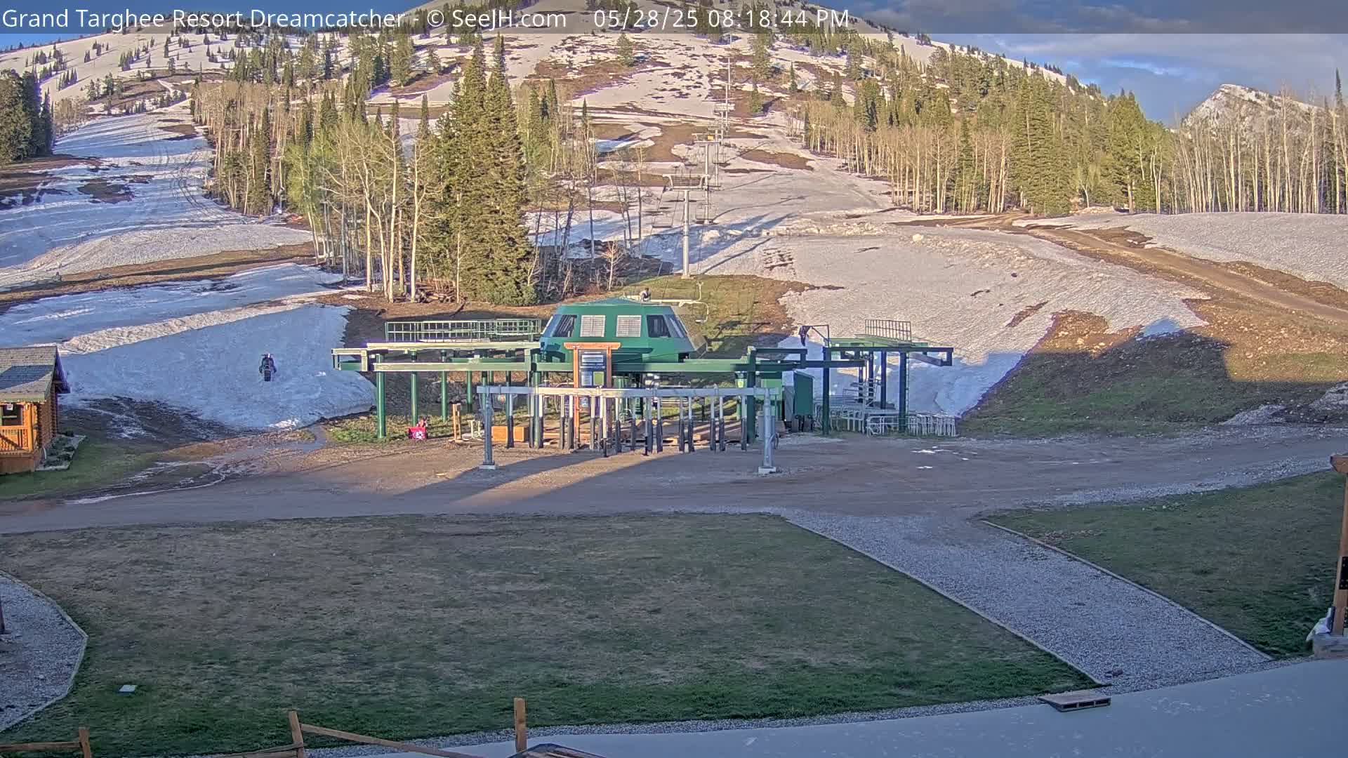 A ski lift sits in a mostly snow-free ski resort area, with patches of snow remaining on the slopes under a partly cloudy sky.