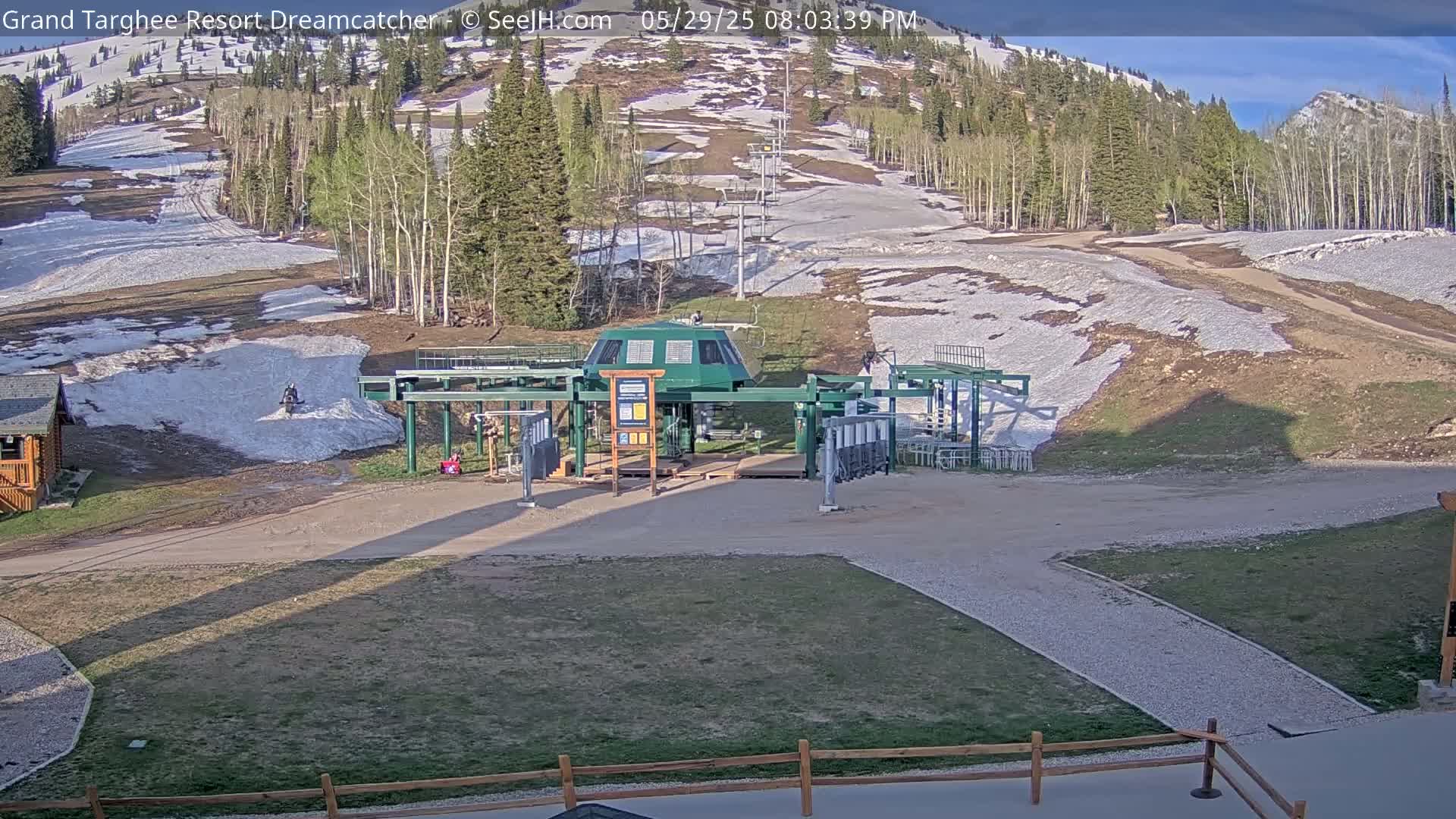 A ski lift base station sits in a grassy area with patches of melting snow on the surrounding hillside under a partly sunny sky.