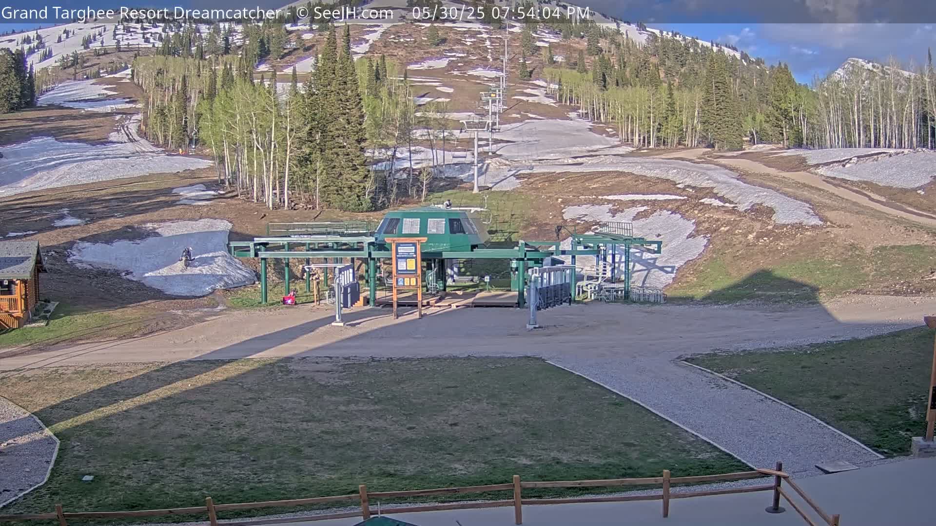A ski lift base station sits in a grassy area with patches of melting snow, backed by a mountainside with trees and more snow.