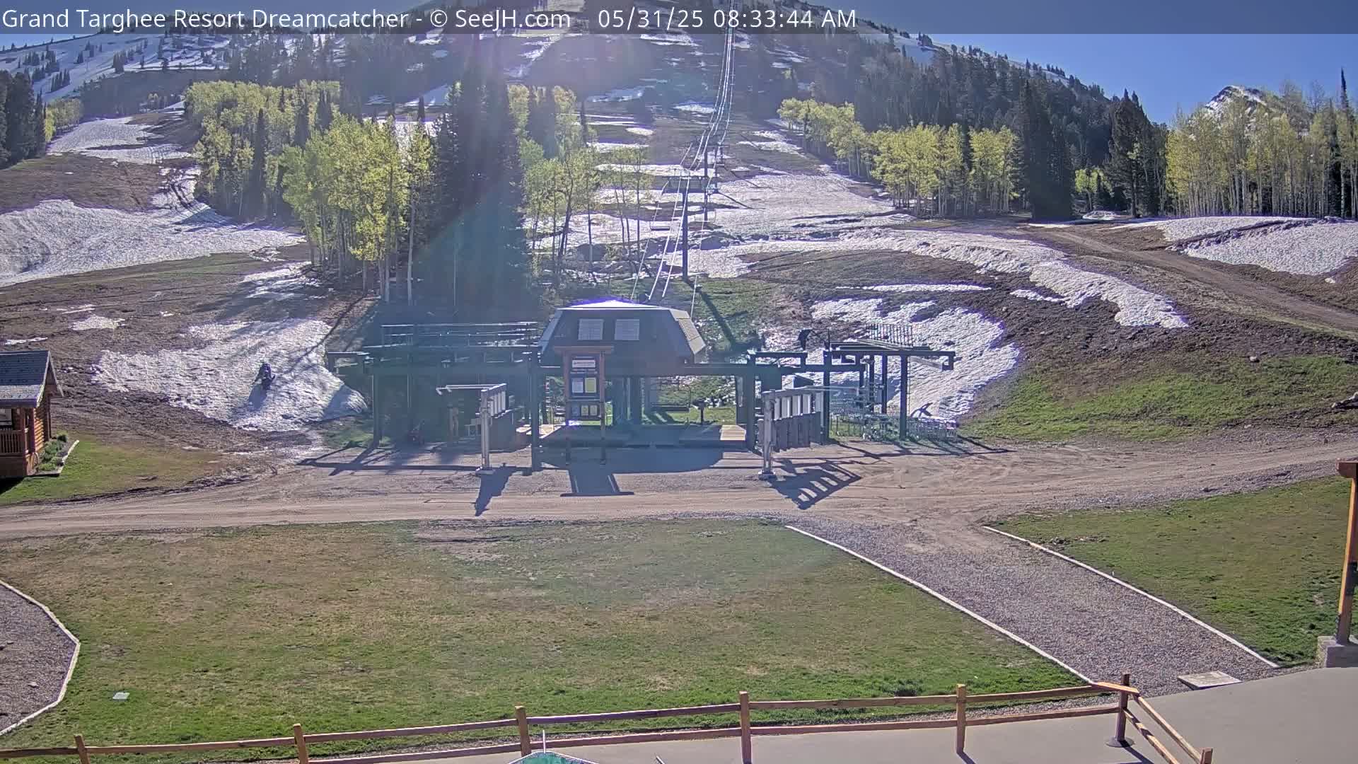 A ski lift base station sits on a grassy area, with patchy snow and dirt visible on the surrounding slopes under a sunny sky.
