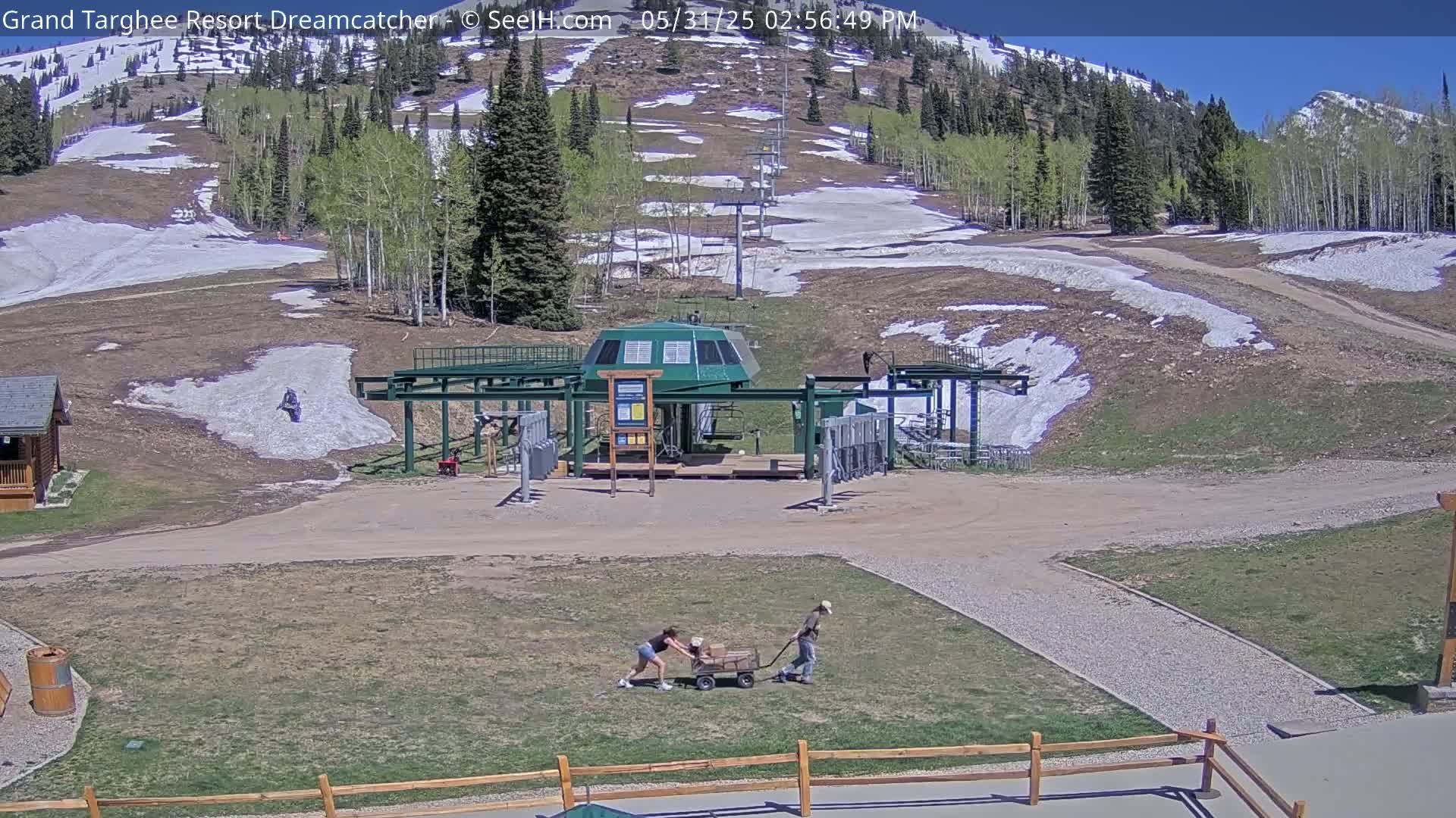 A ski lift base station sits on a grassy area with patches of melting snow on the surrounding hillside under a clear blue sky.