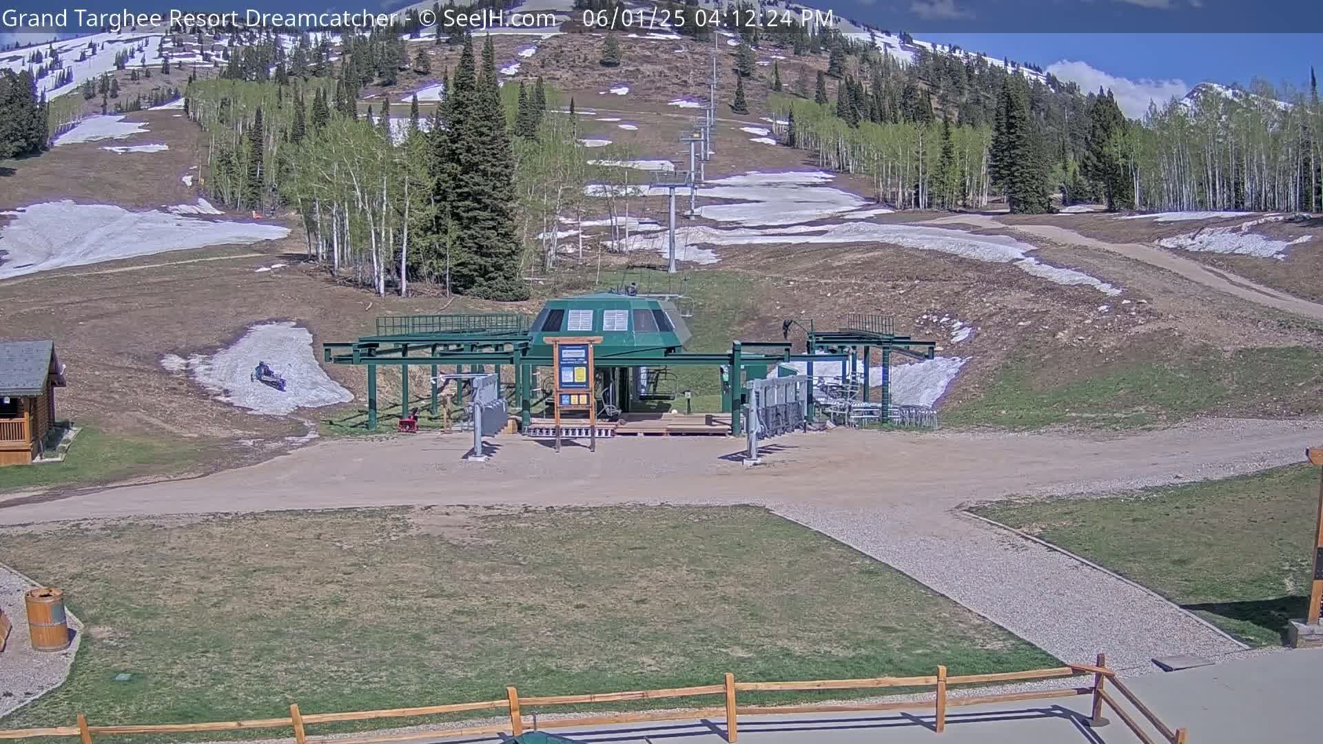 A ski lift base station sits in a grassy area with patchy snow on the surrounding hillside under a mostly sunny sky.
