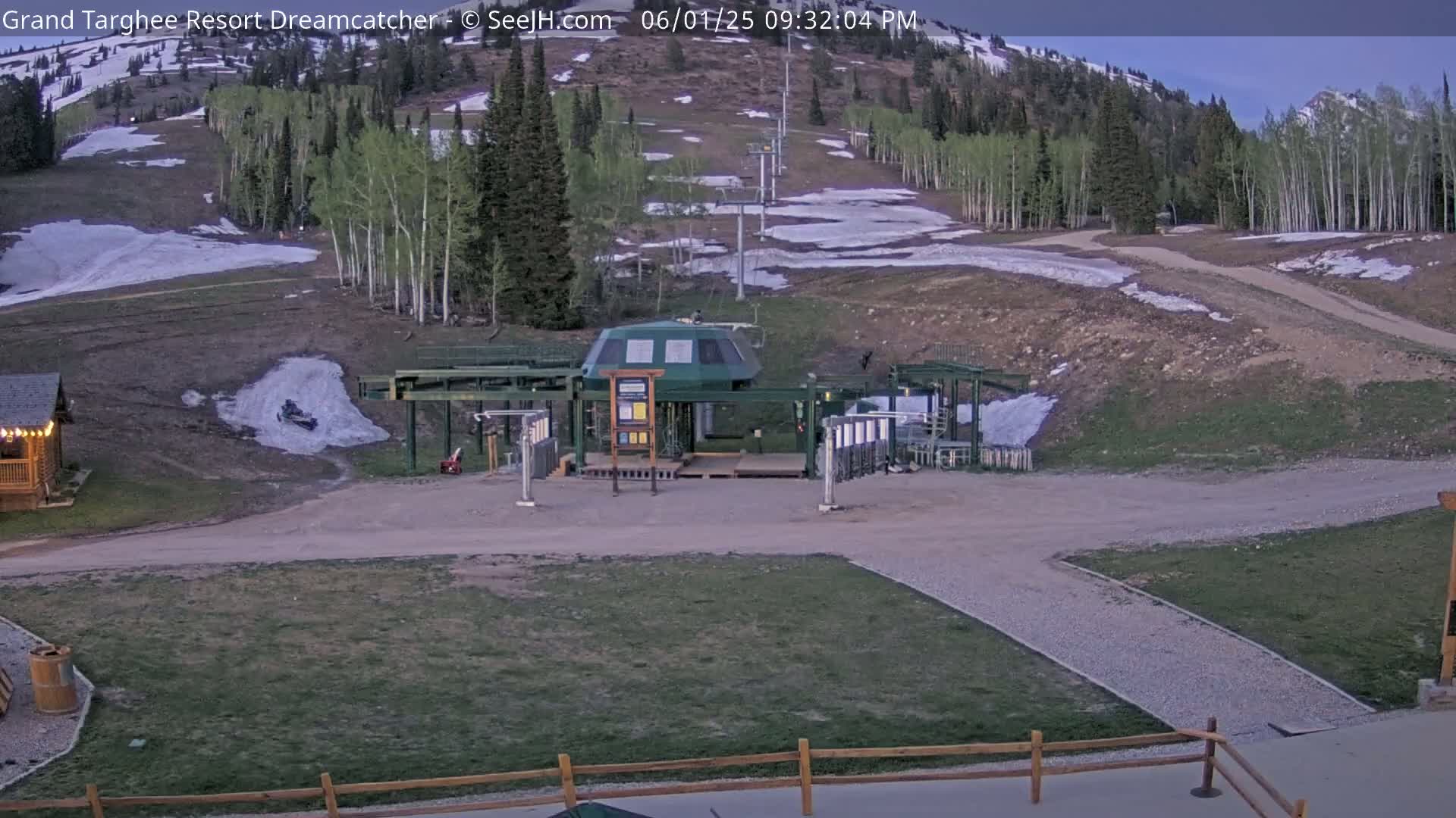 A ski lift base station sits in a grassy area with patches of remaining snow on the surrounding hillside under a twilight sky.