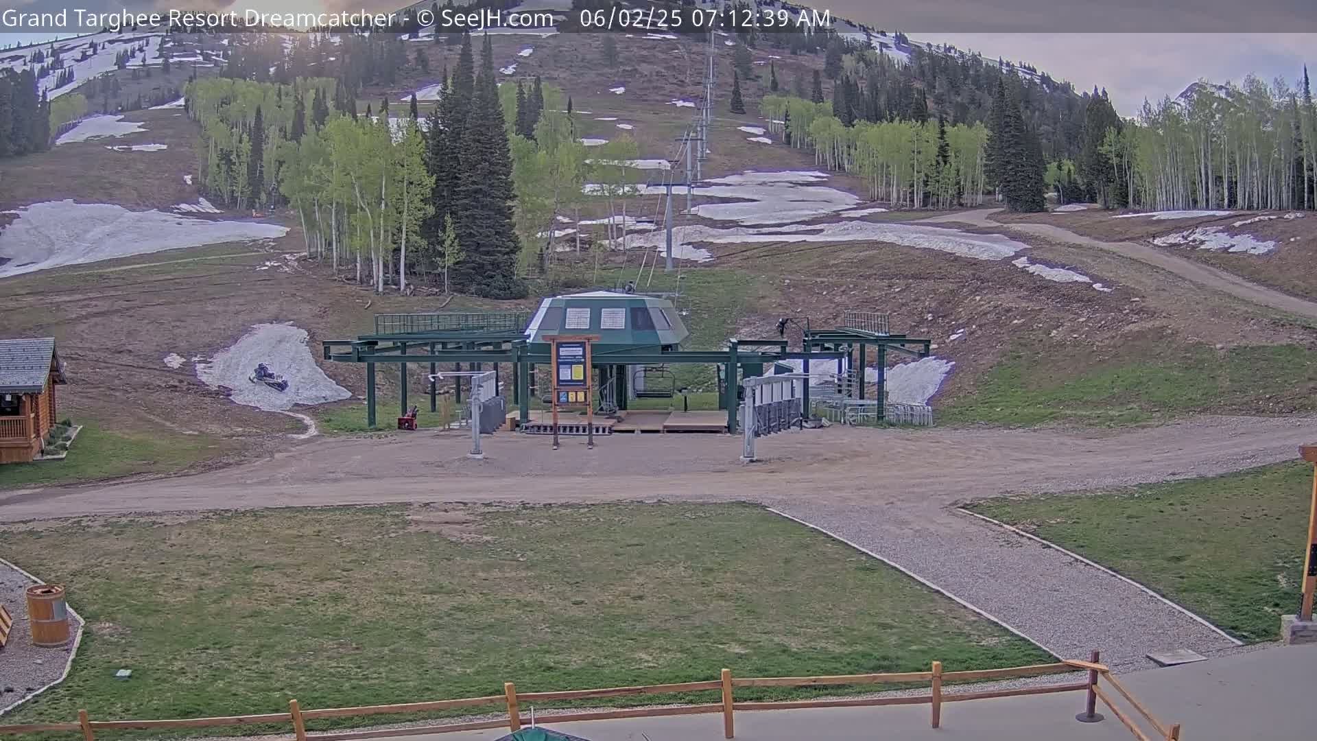 A ski lift base station sits in a mostly grassy area with patches of remaining snow, against a backdrop of a mountainside with sparse trees and melting snow.