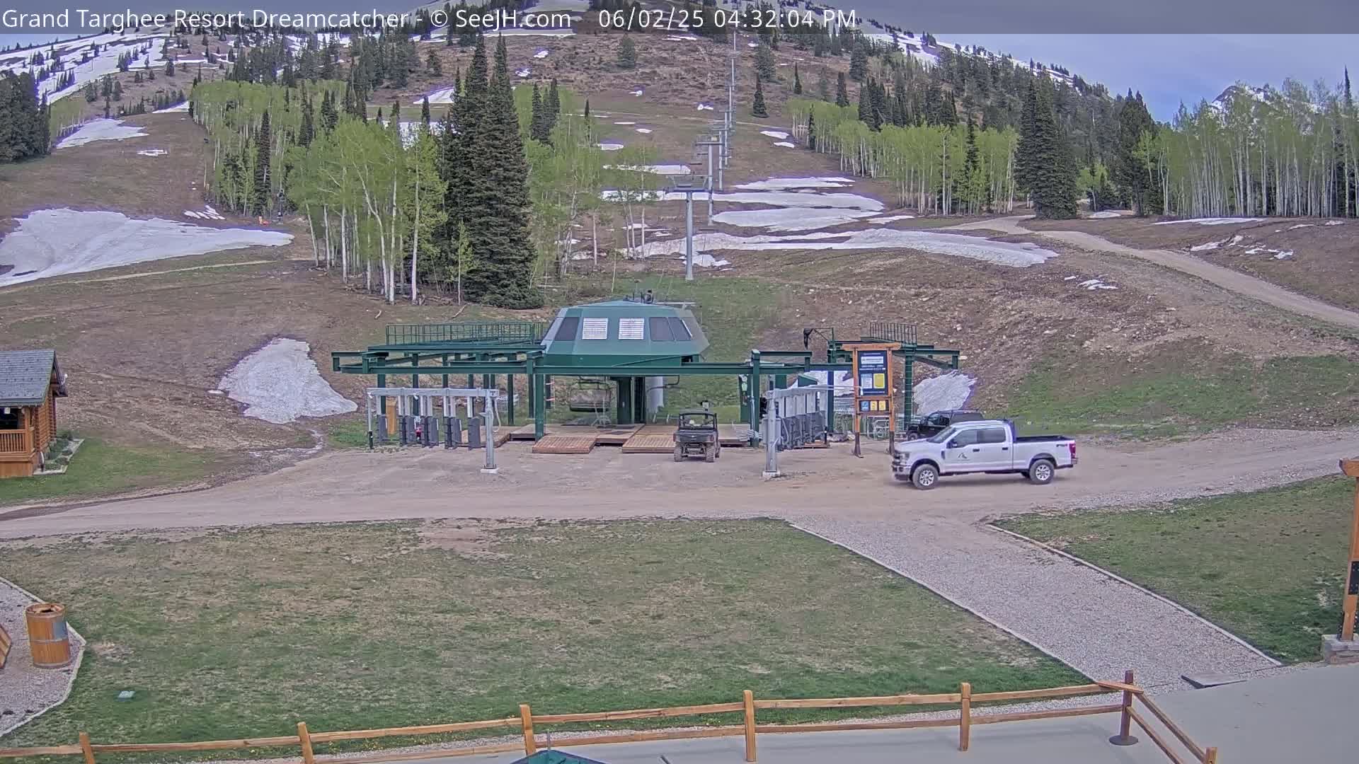 A ski lift base station with a truck and utility vehicle parked nearby sits on a grassy area with patches of melting snow in the background under a partly cloudy sky.