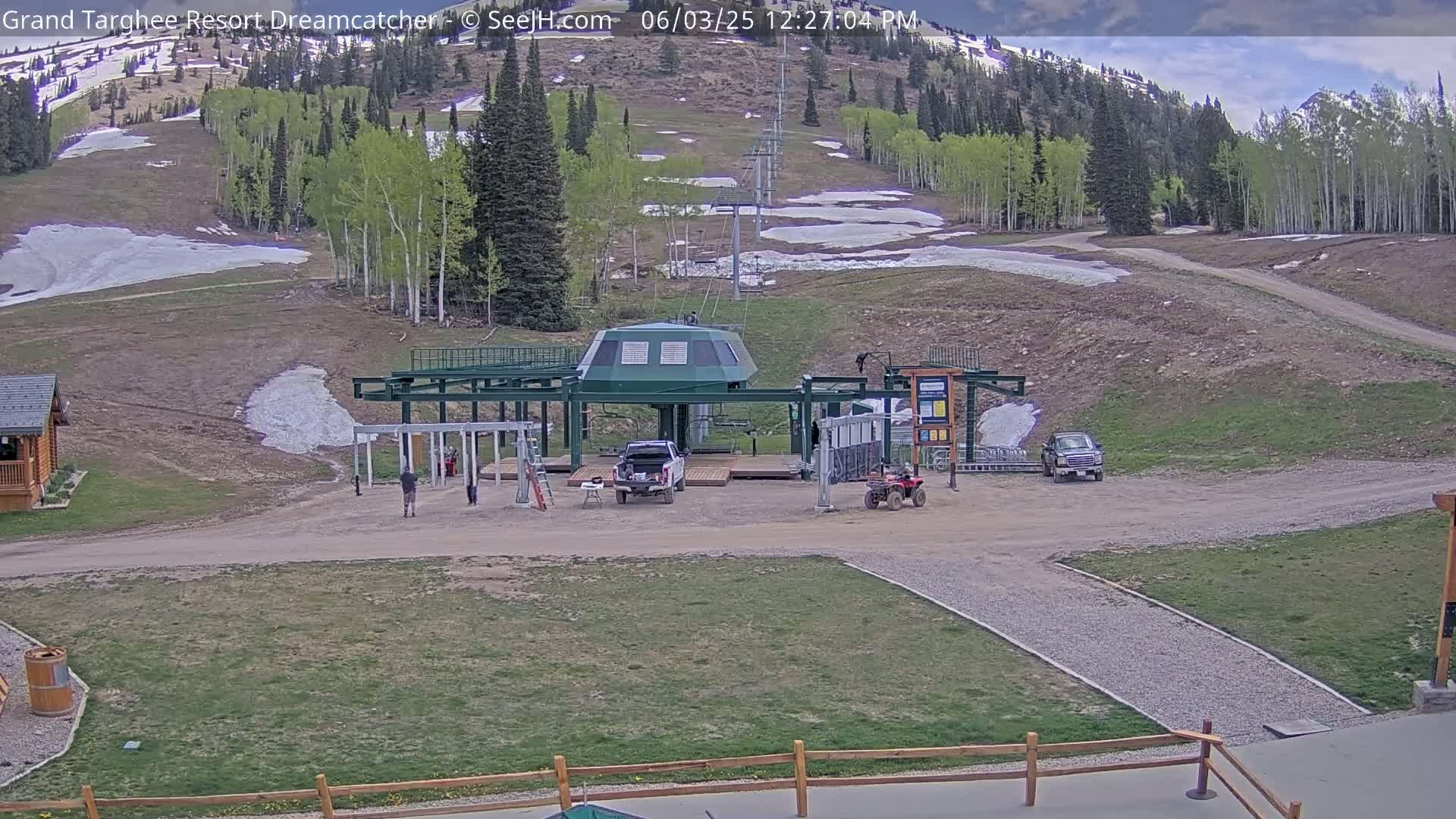 A ski lift base station is situated on a grassy area with patches of snow, partially cloudy skies, and a mountainside visible in the background.
