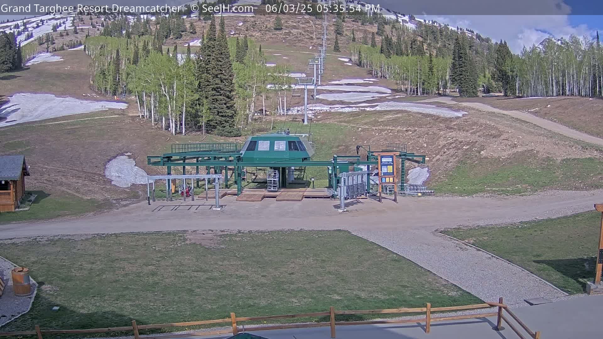 A ski lift base station sits on a grassy area with a partially snow-covered hillside in the background under partly sunny skies.