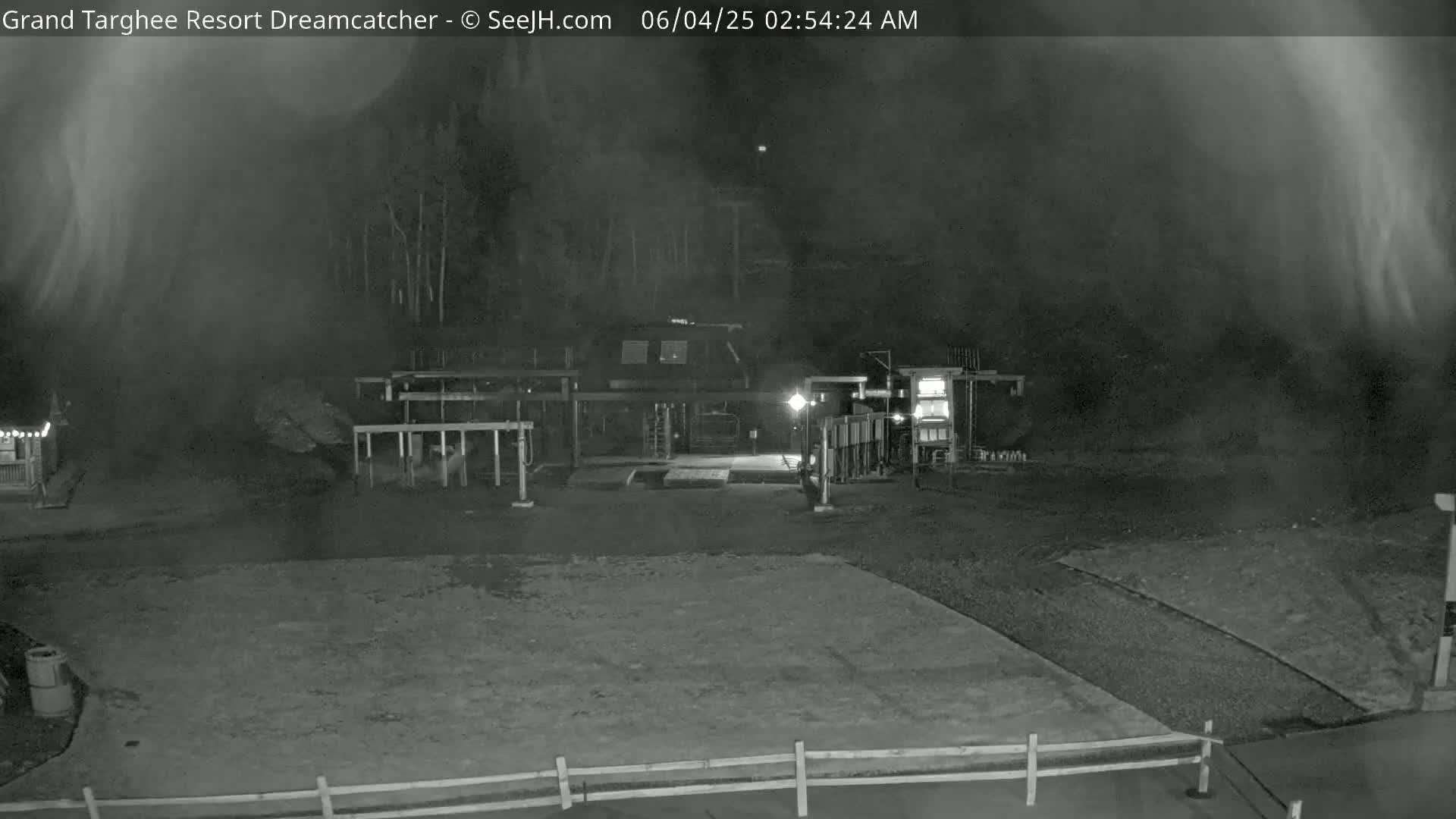 A nighttime, hazy view of a mostly empty, paved area in front of a building with some structures and a low fence.