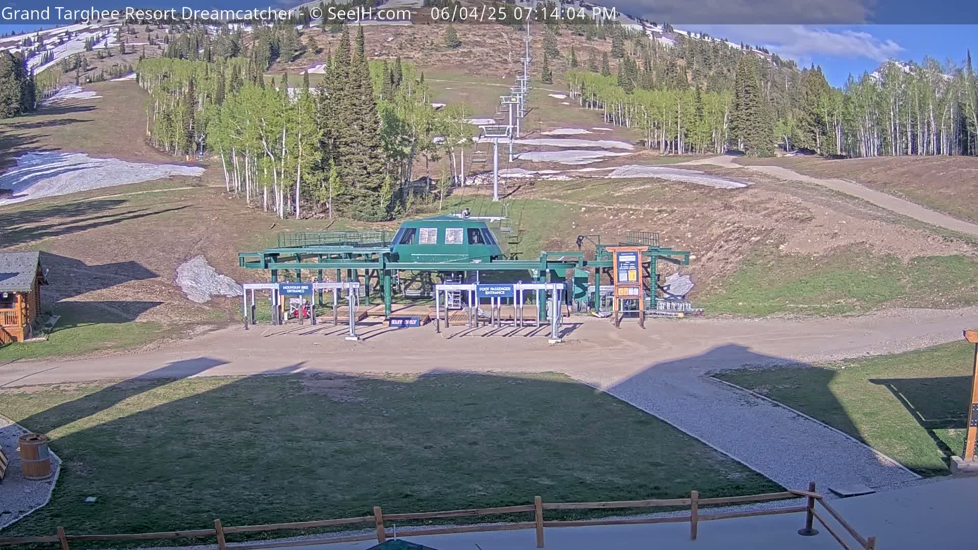 A ski lift base station sits in a grassy area with patches of snow remaining on the hillside under a mostly sunny sky.