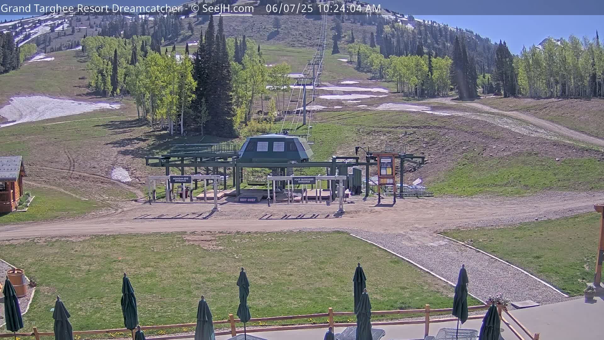 A chairlift base station sits in a grassy area with a dirt path leading to a mountainside with patches of remaining snow under a sunny sky.