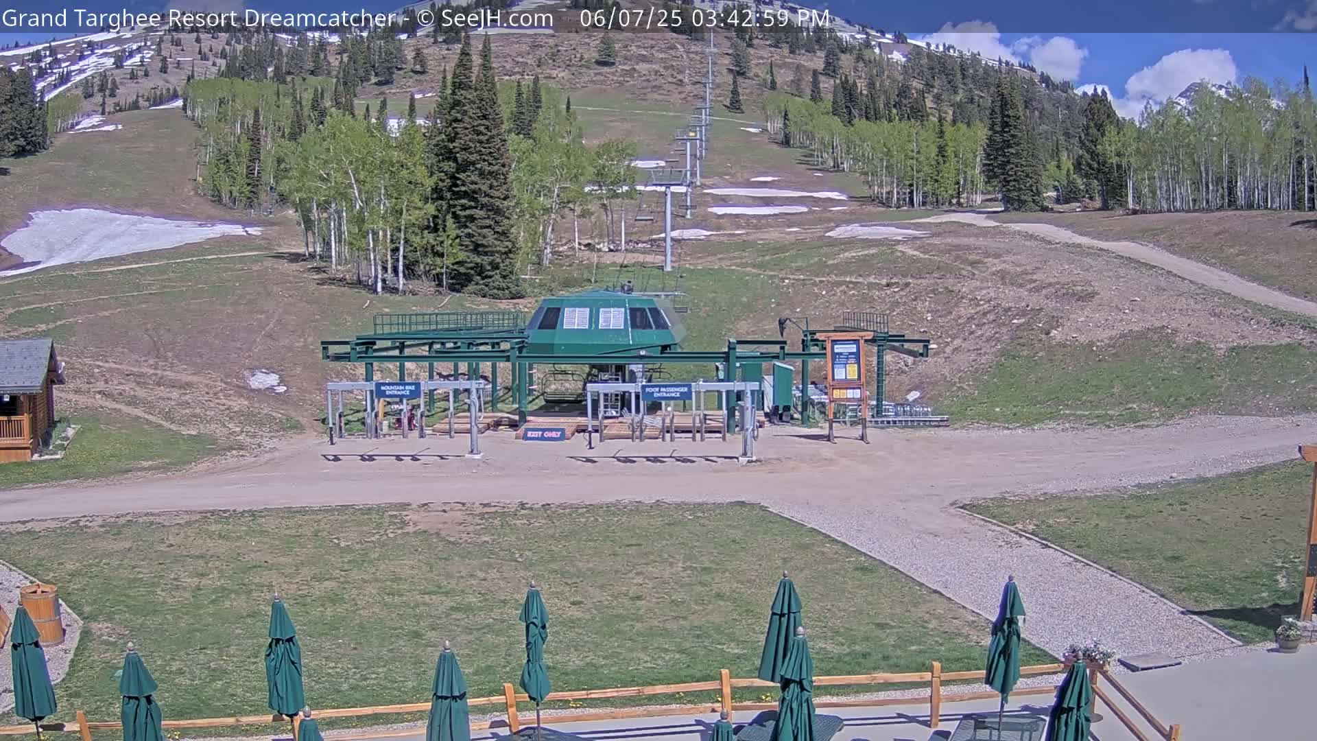A ski lift base station sits on a grassy area under a mostly sunny sky, with a mountainside partially covered in snow visible in the background.