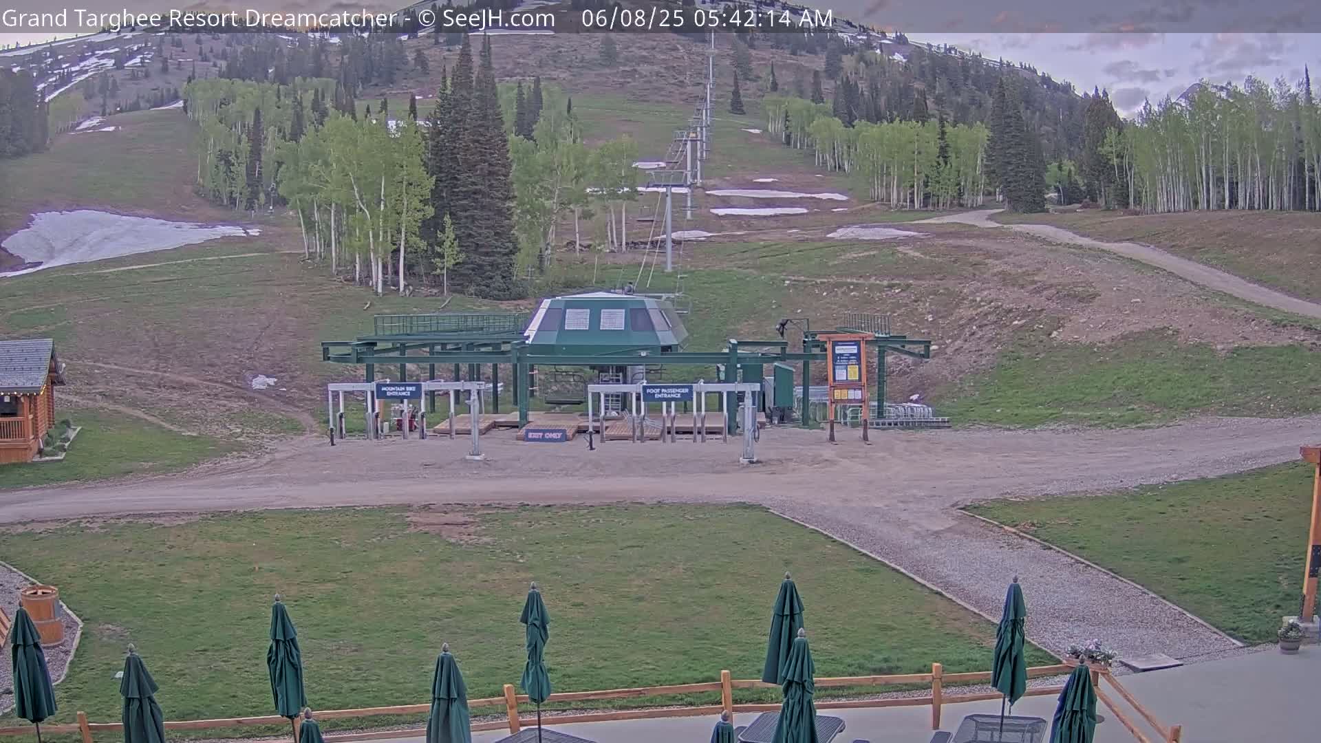 A ski lift base station sits at the bottom of a grassy hill with a few patches of remaining snow under a partly cloudy sky.