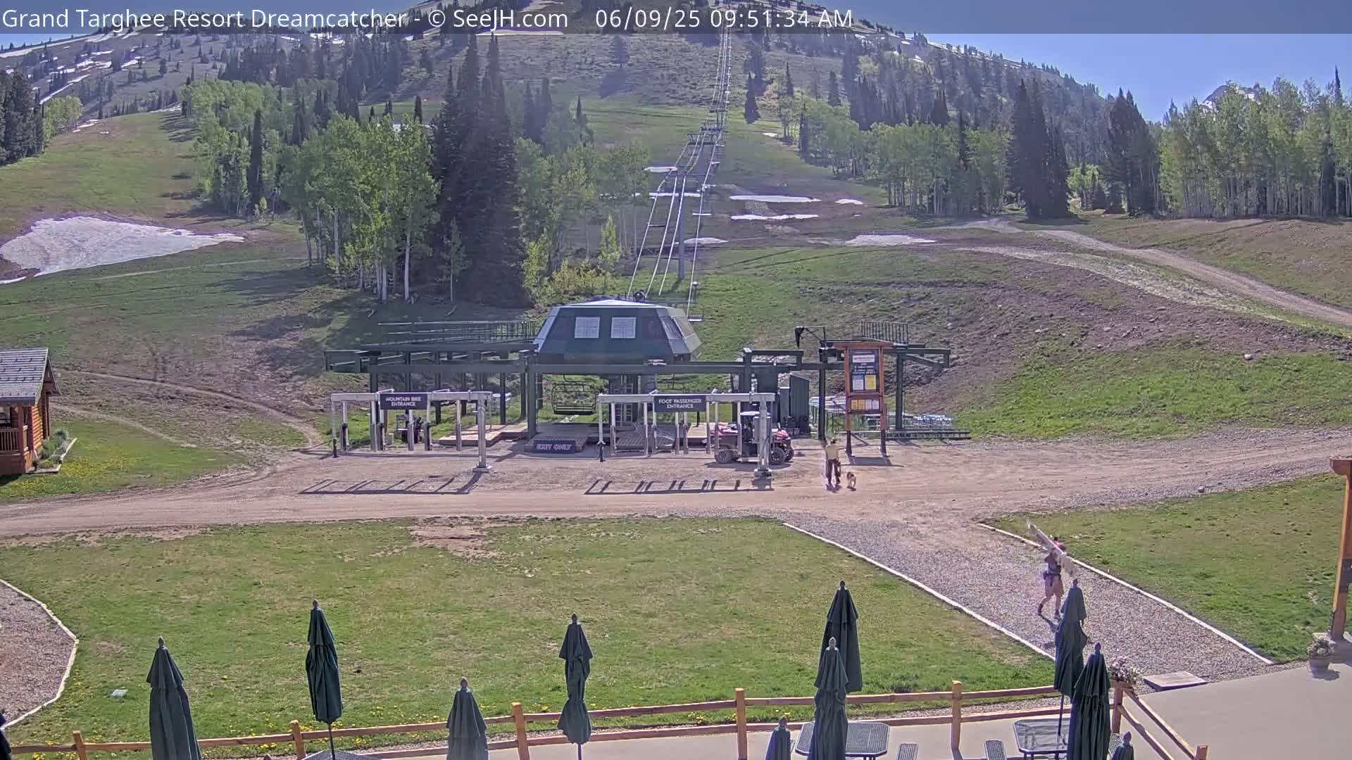 A mountain ski lift base area on a sunny day, with some remaining snow patches visible on the slopes, features a few people and dogs near the lift, and patio umbrellas in the foreground.