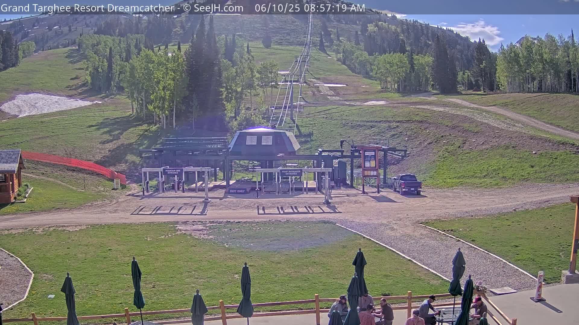 A ski lift base area with a mostly sunny sky and a few patchy clouds, showing a grassy hill, a parking lot with a vehicle, and people sitting under umbrellas at outdoor seating.