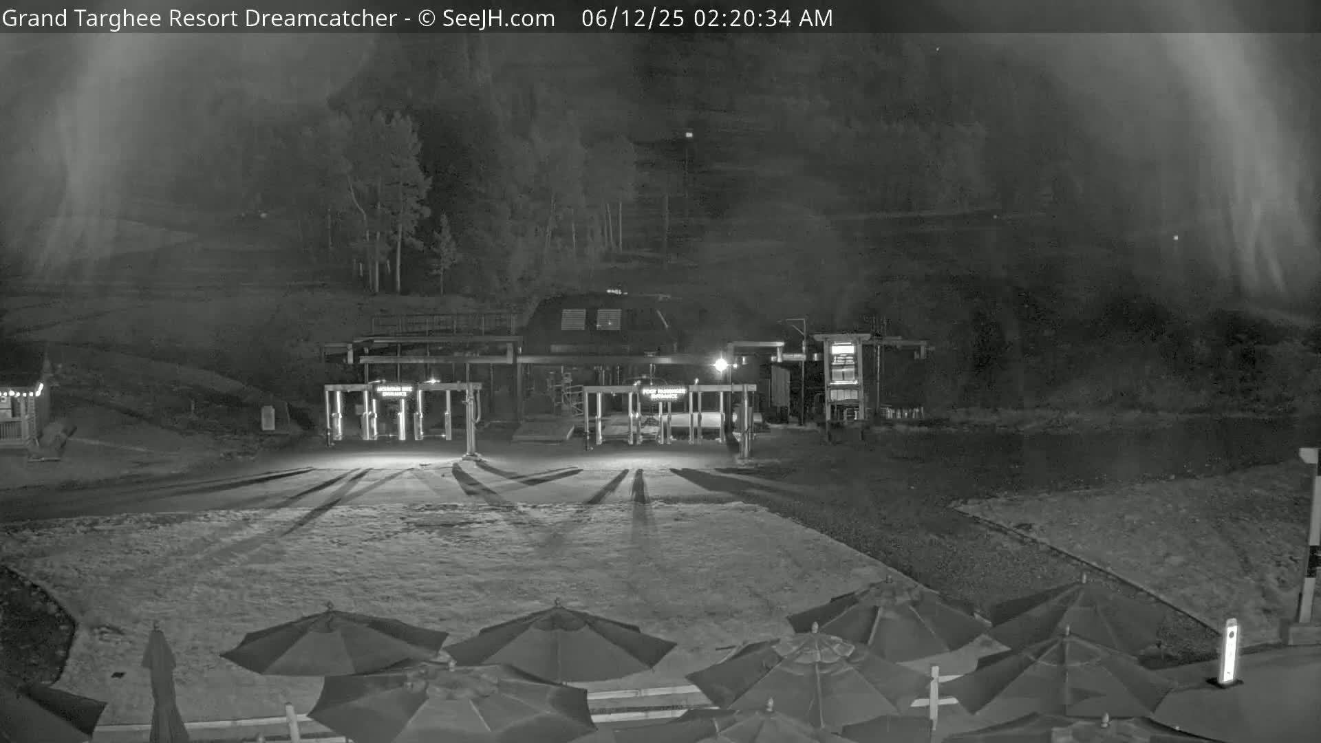 A nighttime, foggy view of a ski resort entrance with closed gates, several closed umbrellas, and a building in the background.