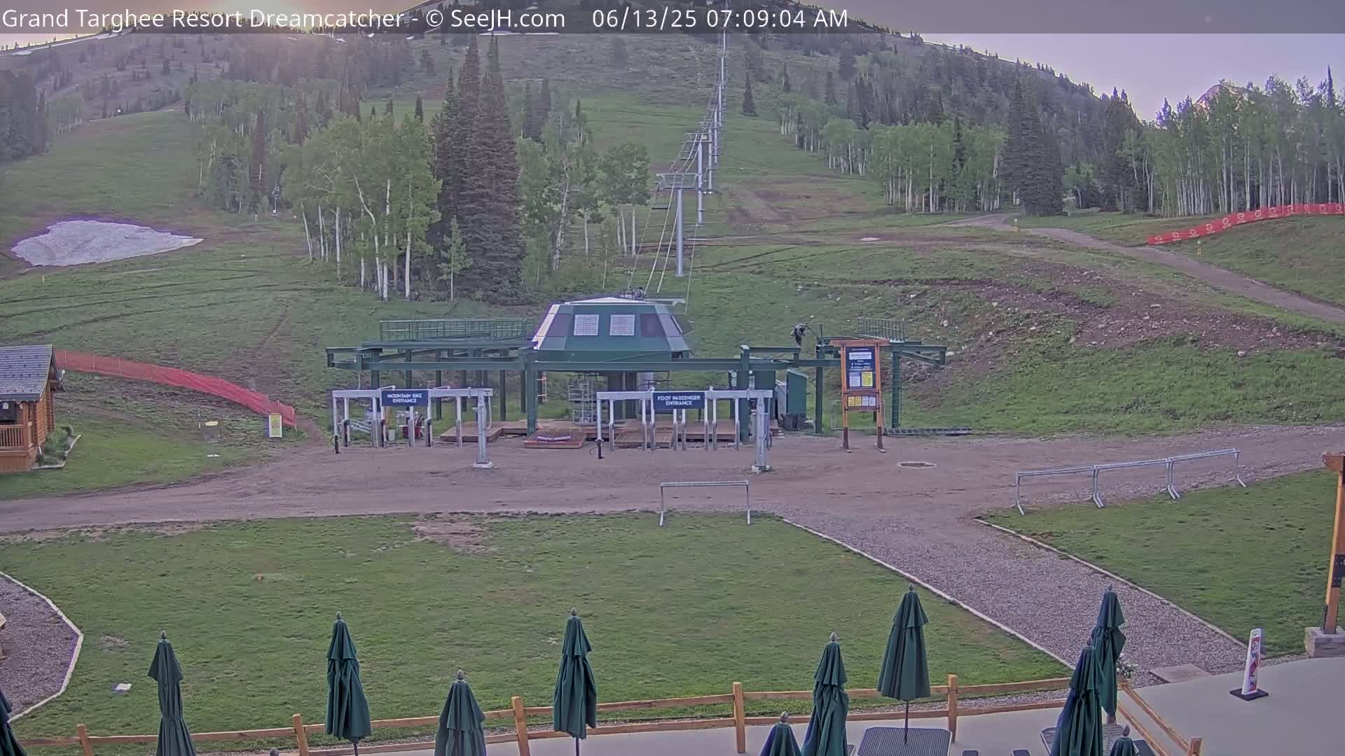A mountainside chairlift base station with a grassy area and several closed dark-green umbrellas in the foreground, under a mostly clear sky.