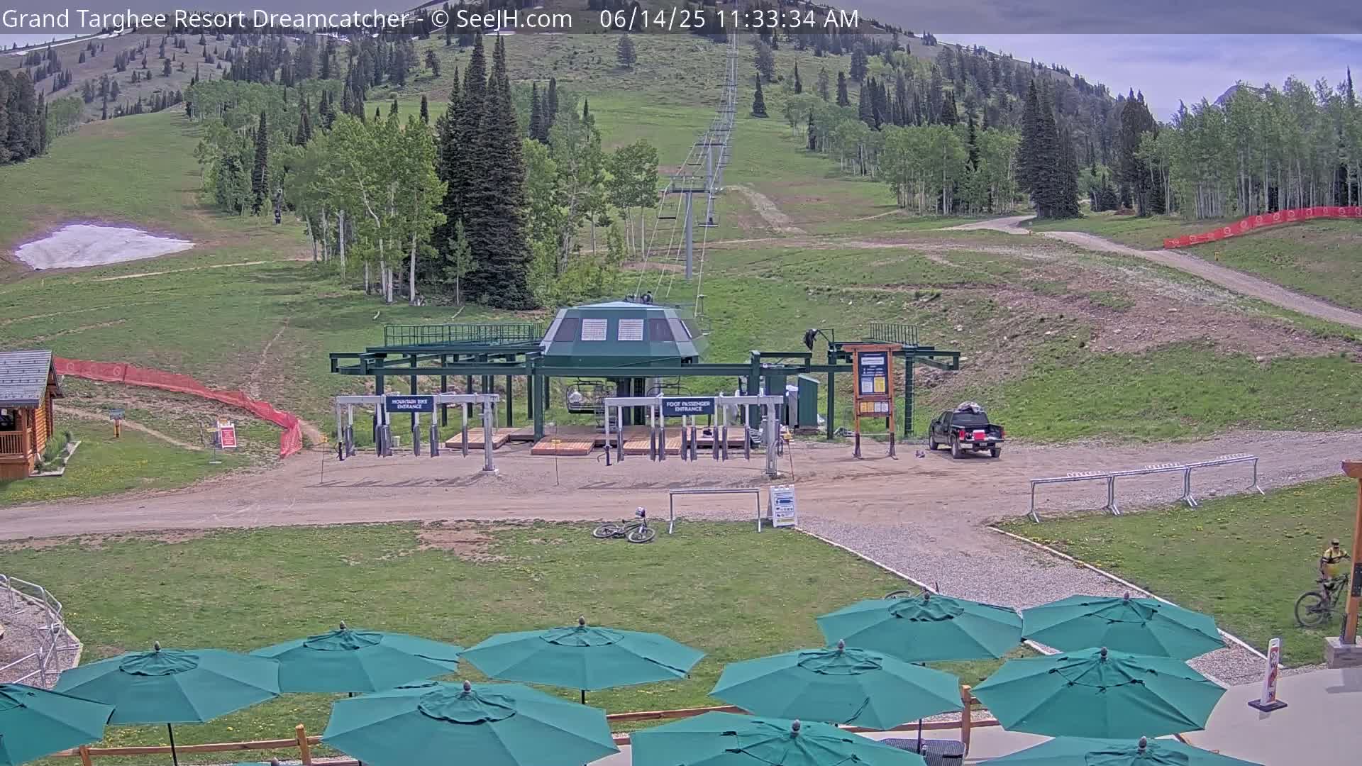 A mostly sunny mountainside scene shows a chairlift base station, a few parked vehicles, some green patio umbrellas, and a mountain bike.