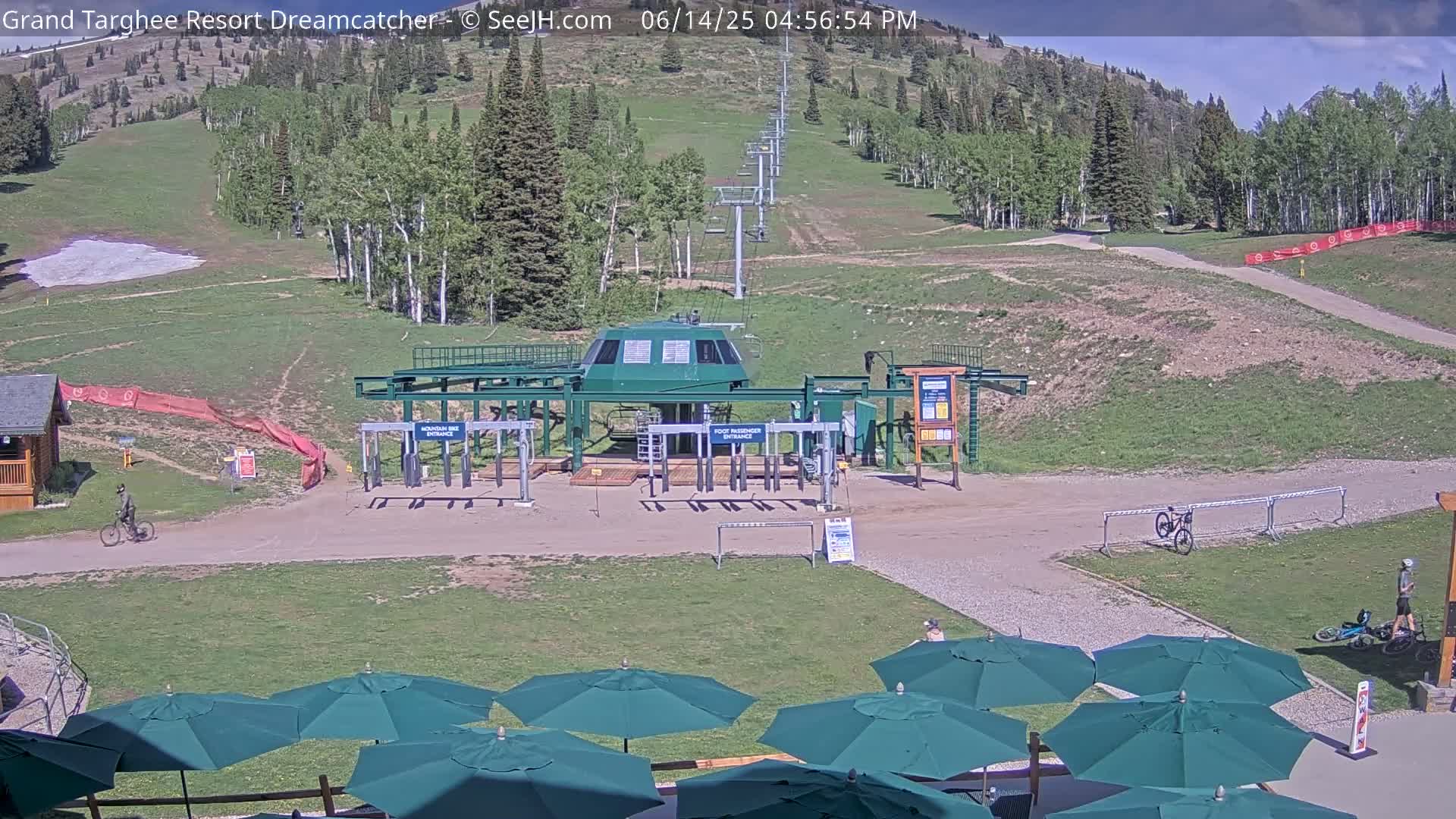 A mostly sunny mountainside scene shows a chairlift base station, a few parked vehicles, some green patio umbrellas, and a mountain bike.