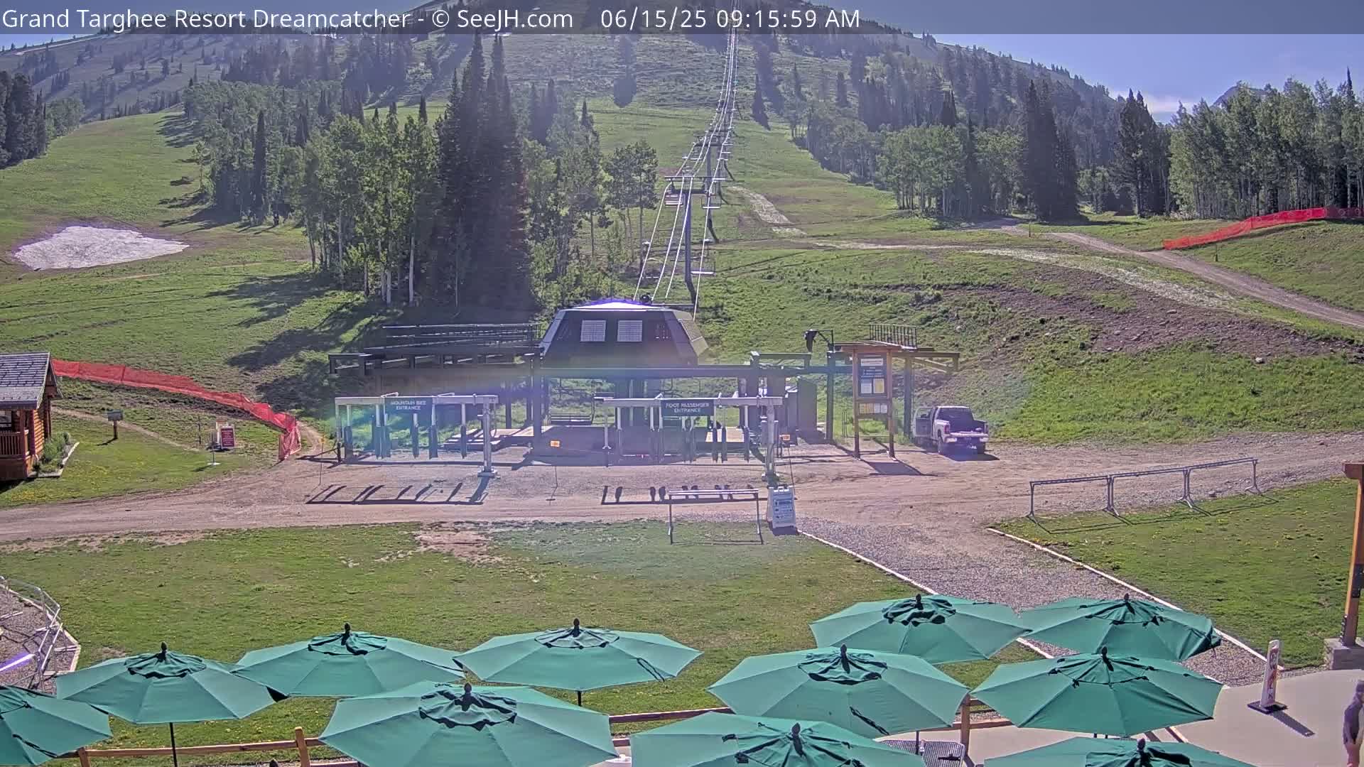 A chairlift base station sits on a grassy hillside under a sunny sky, with several green umbrellas arranged in the foreground.