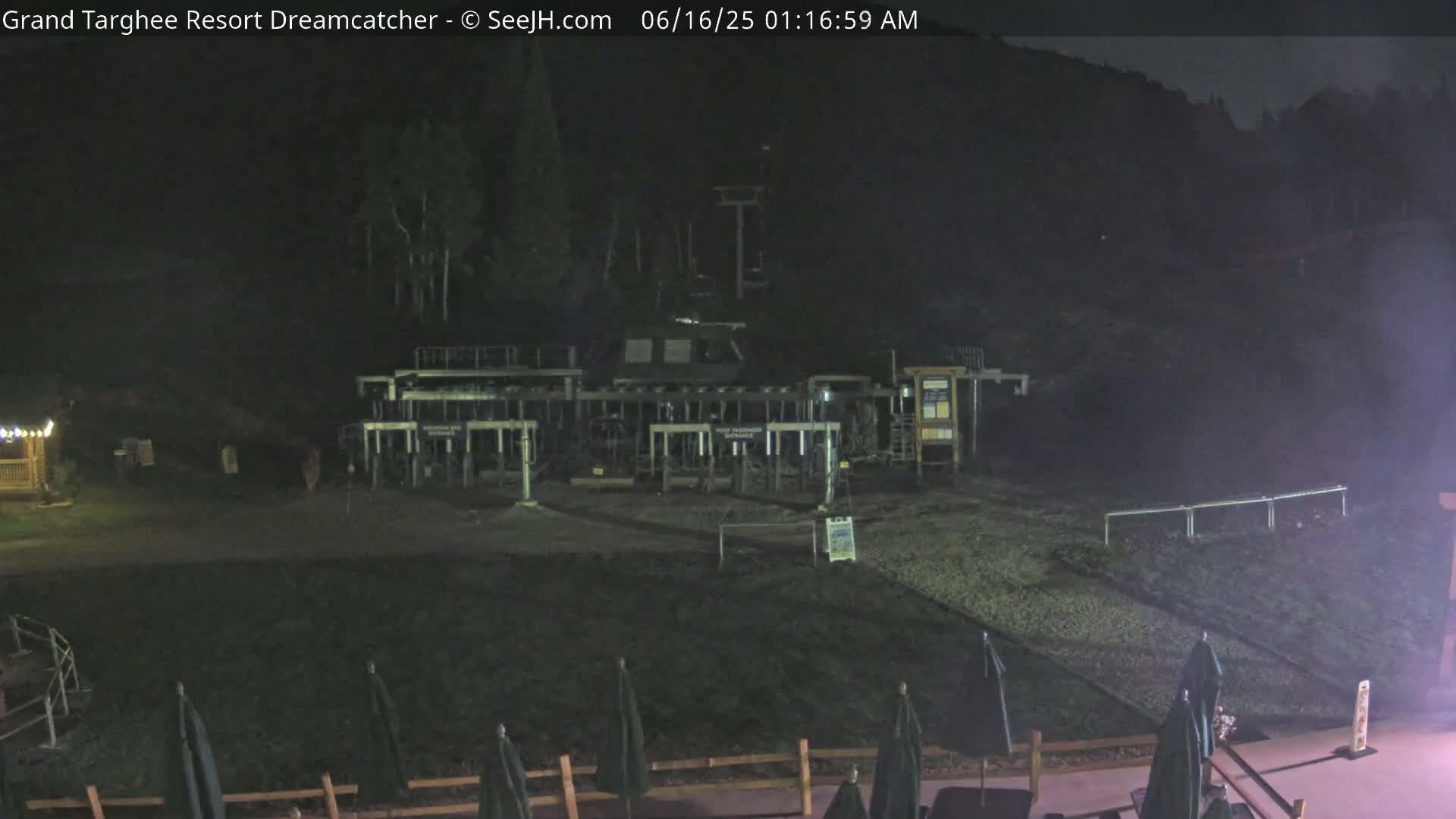 A nighttime view of a ski lift base area with several closed gondolas, a dark hillside in the background, and a few closed umbrellas in the foreground.