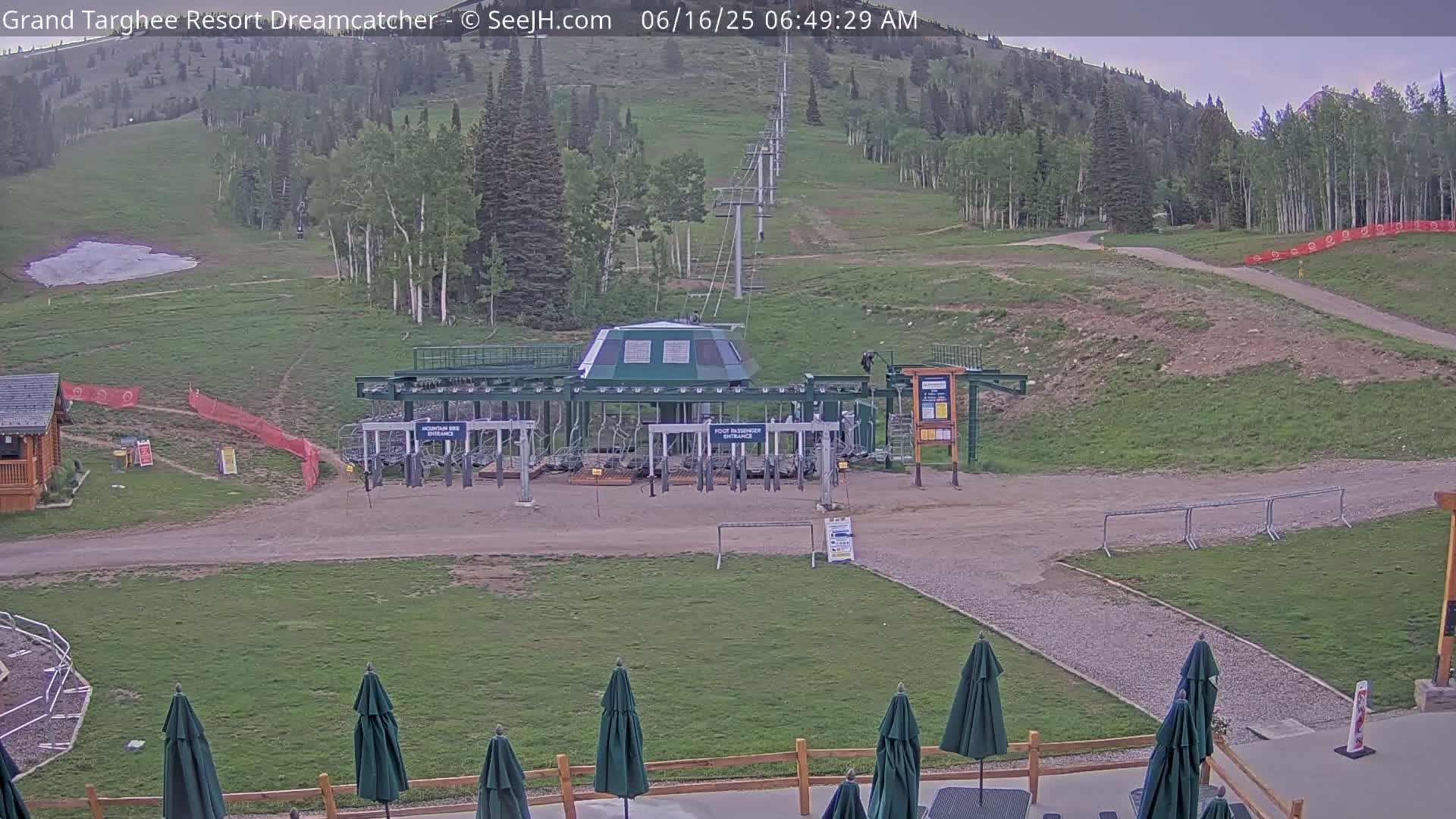 A chairlift base station sits on a grassy area with a mountainside covered in green trees in the background under a partly cloudy sky.