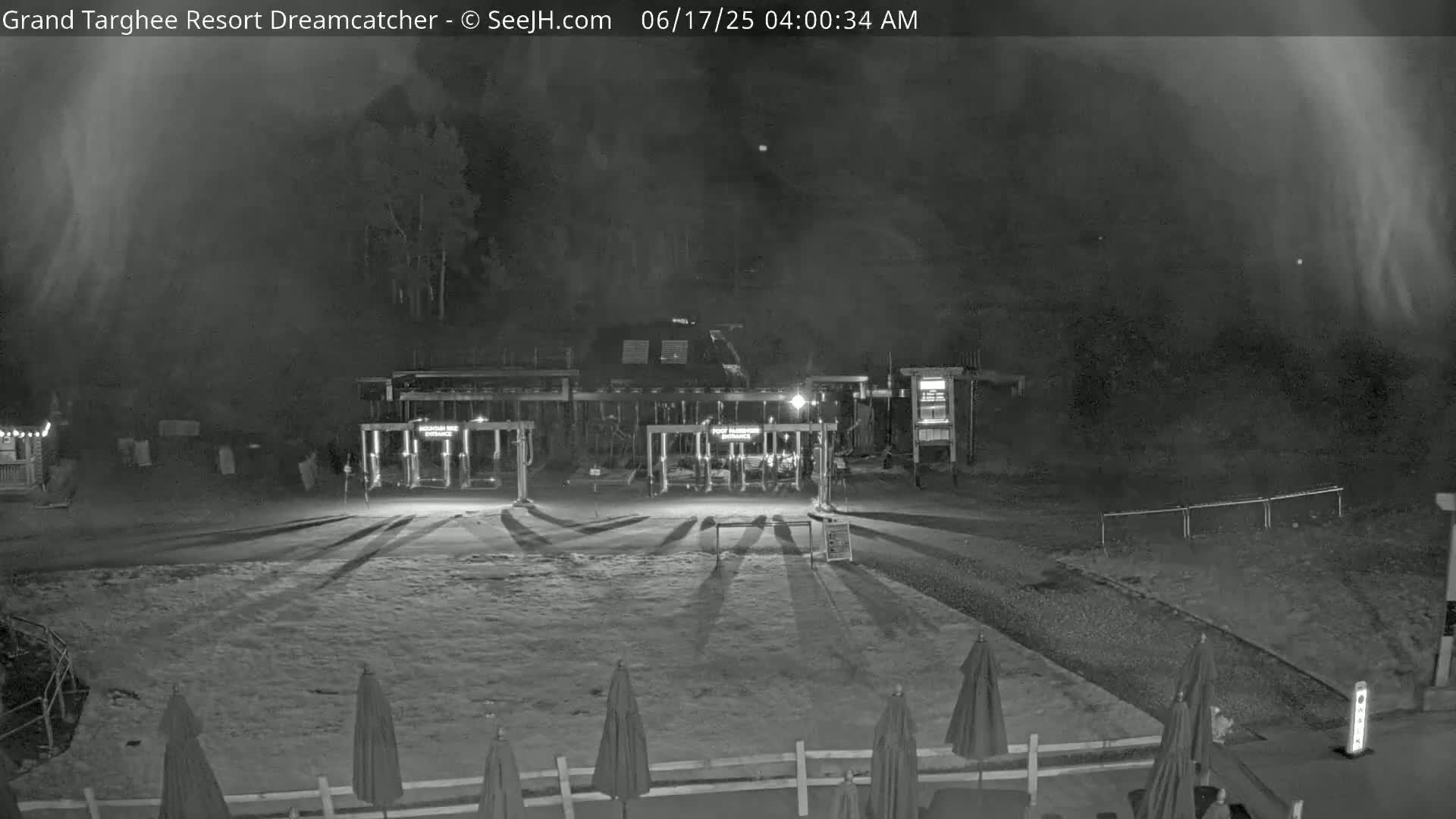 A nighttime, hazy view of a ski resort entrance with closed gates, several closed umbrellas, and long shadows cast on the snow-covered ground.