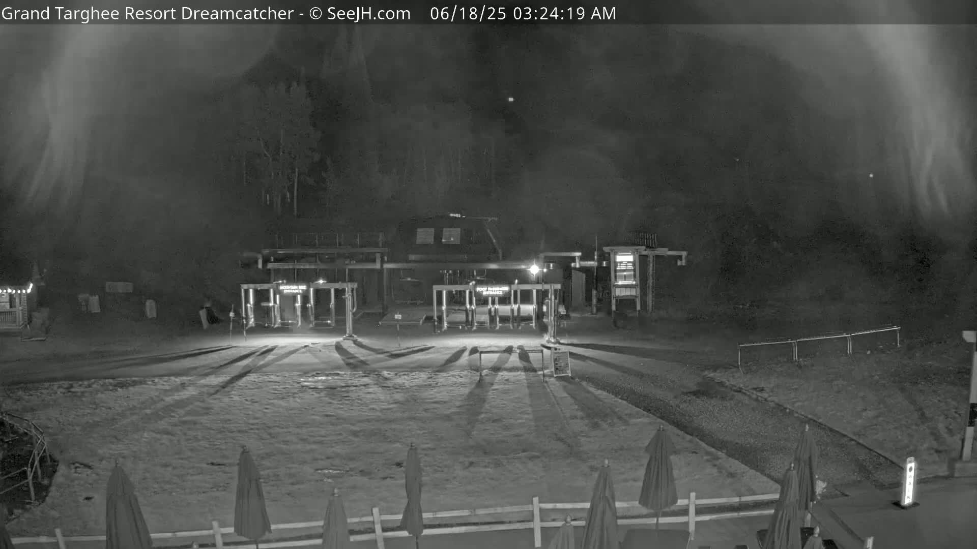 A nighttime, snow-covered scene shows a resort entrance with closed gates and several closed umbrellas in the foreground under a hazy sky.