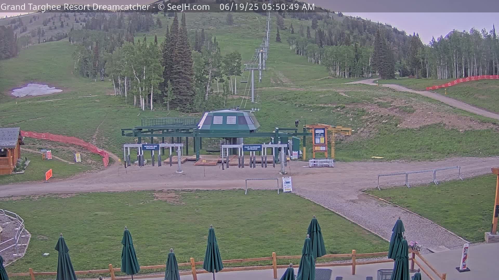 A ski lift base station sits on a grassy hill under a mostly clear sky, with several closed umbrellas and outdoor furniture visible in the foreground.